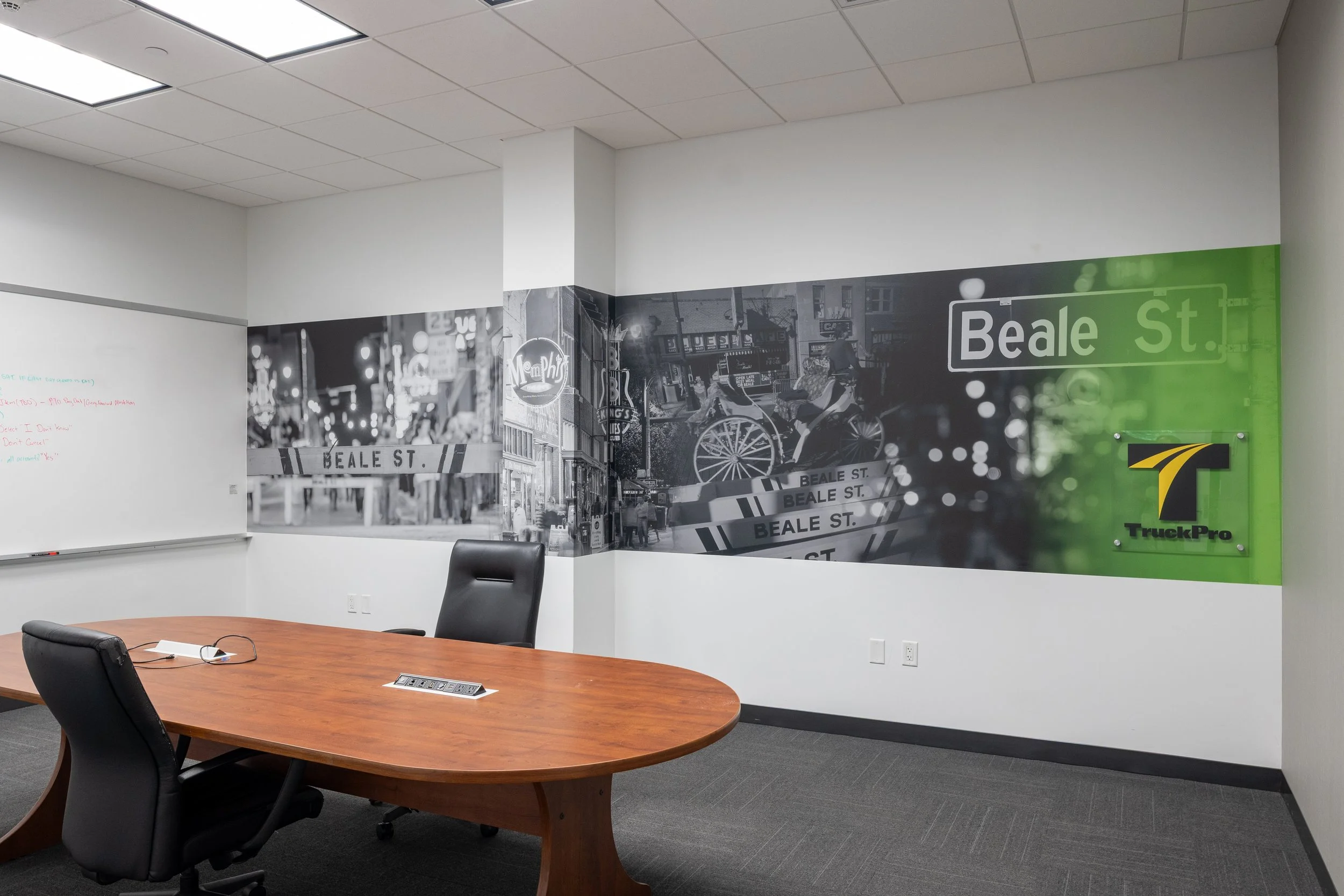 An office conference room with a wooden table and black chairs, a wall with urban street art, and a whiteboard to the left.