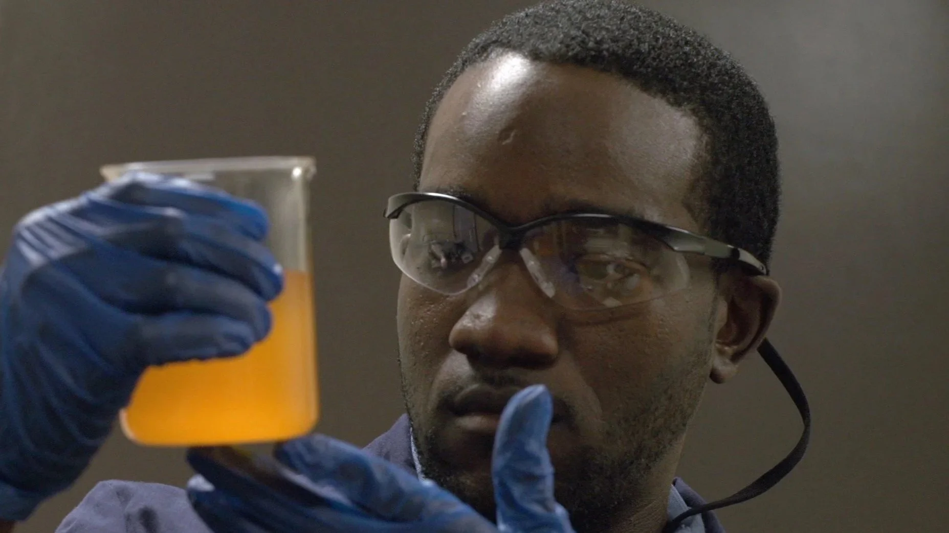 A scientist wearing safety glasses and blue gloves examining a glass of orange liquid.