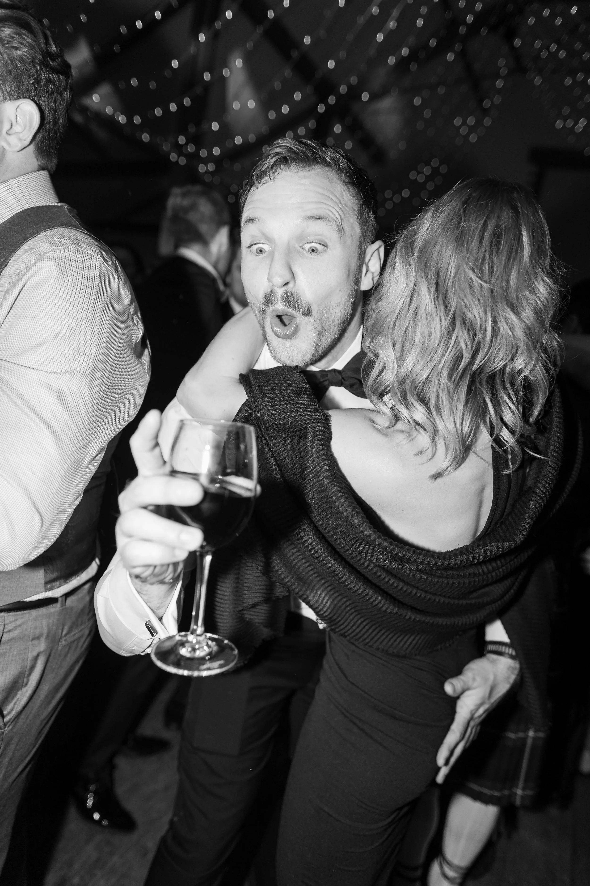 Man in tuxedo with surprised expression holding glass of wine at party.