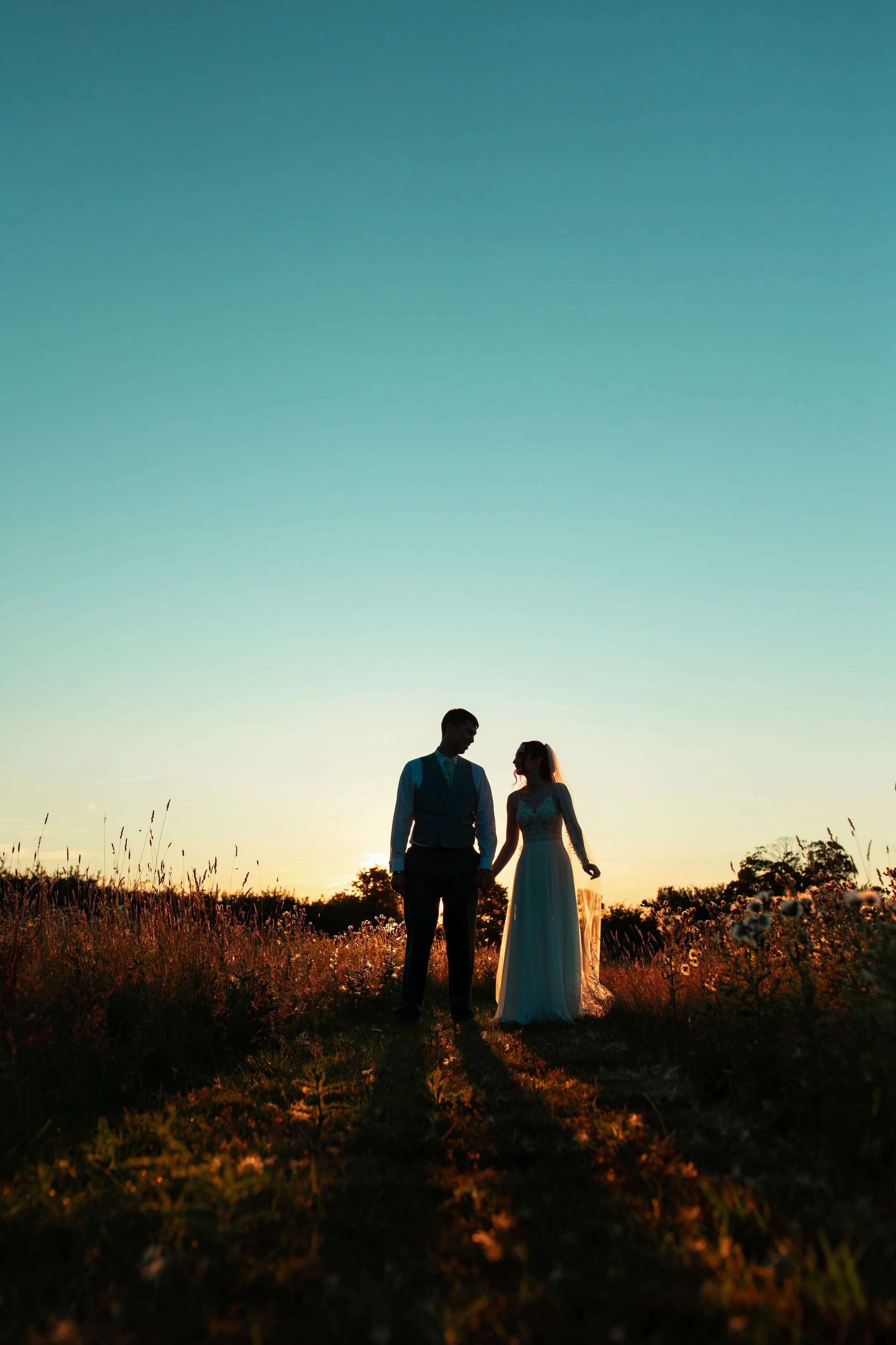 A couple, a man and a woman in wedding attire, holding hands in a field during sunset with a clear sky.