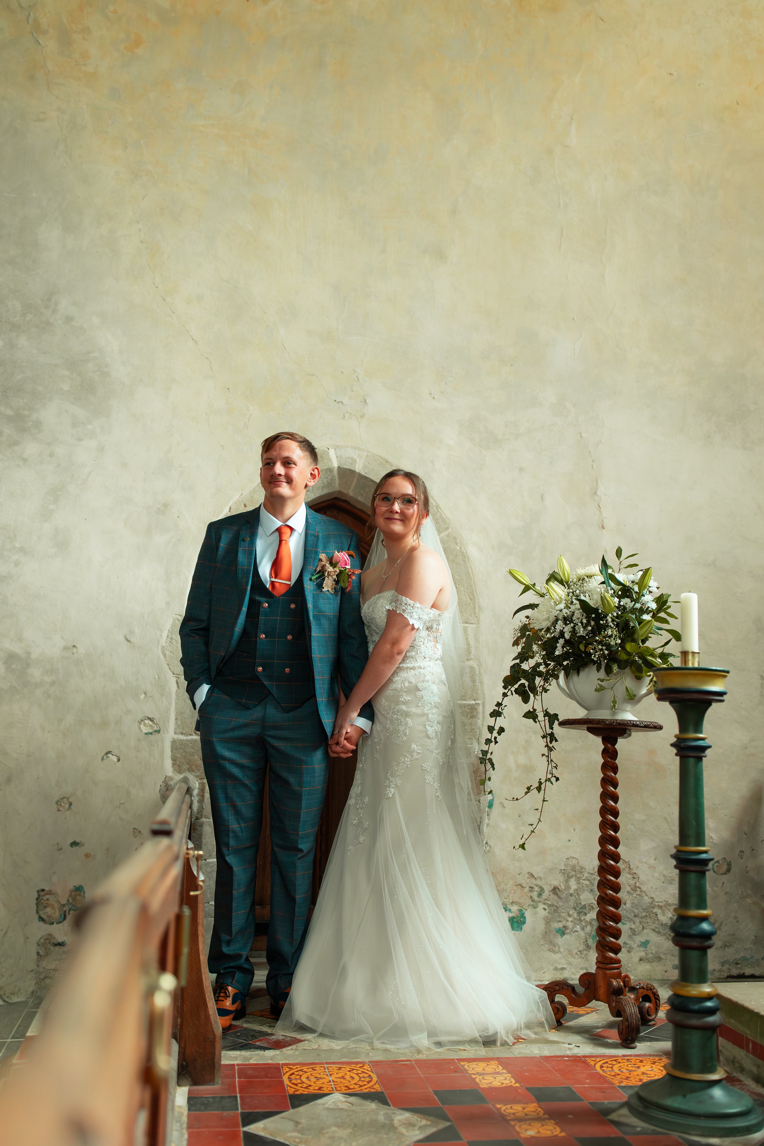 A bride and groom standing hand in hand during their wedding ceremony, indoors with a rustic stone wall background and a floral arrangement on a stand.