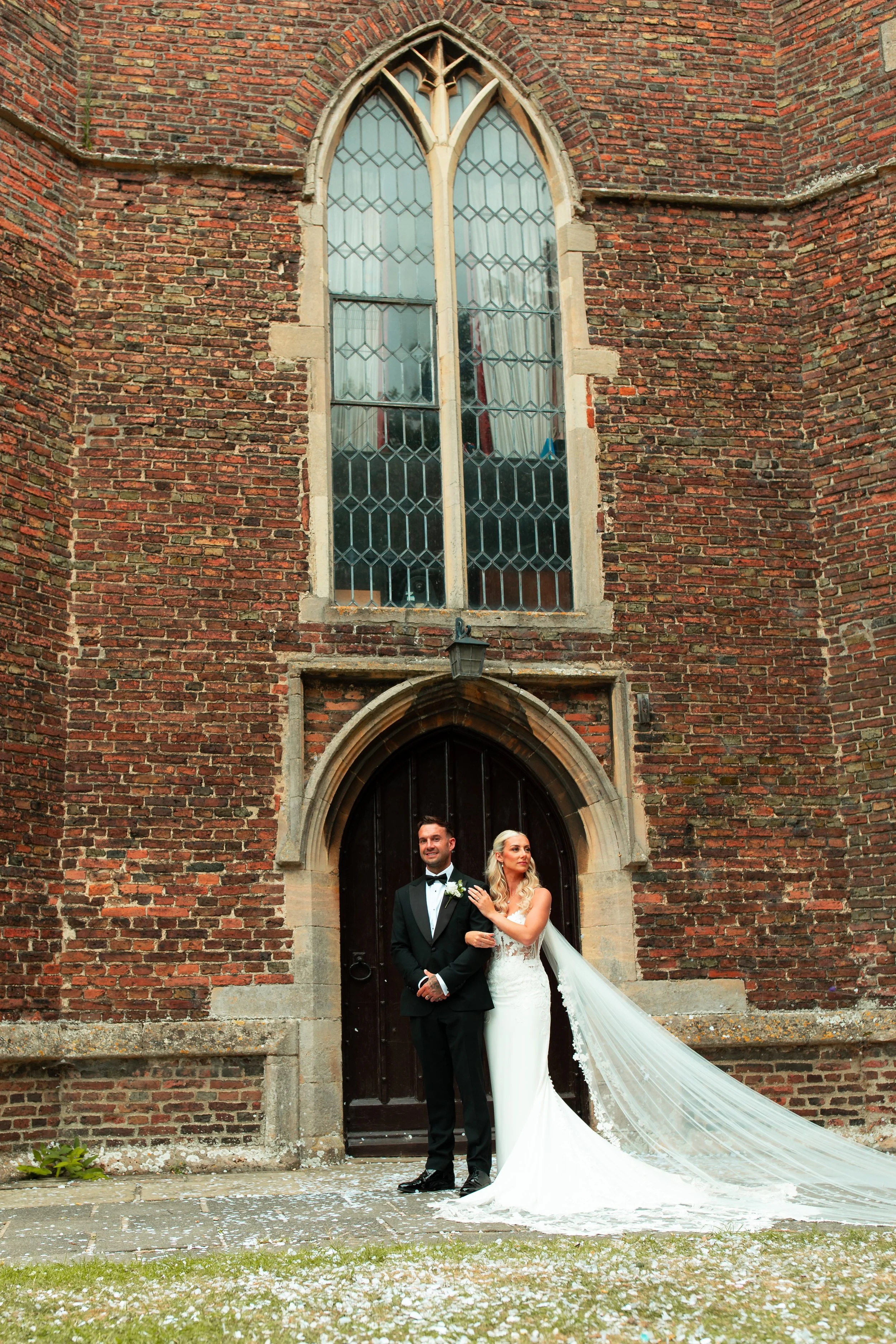 Bride in a white wedding gown with a long train and veil, and groom in a black tuxedo, standing in front of a brick church with a large arched wooden door and stained glass window above.