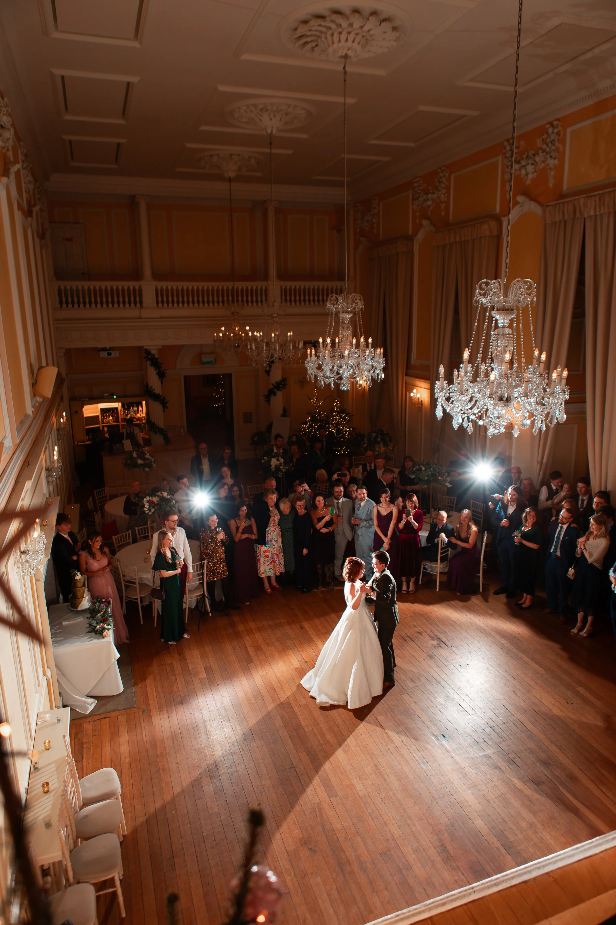 A bride and groom dancing in the center of a grand ballroom with chandeliers, surrounded by guests watching at a wedding reception.