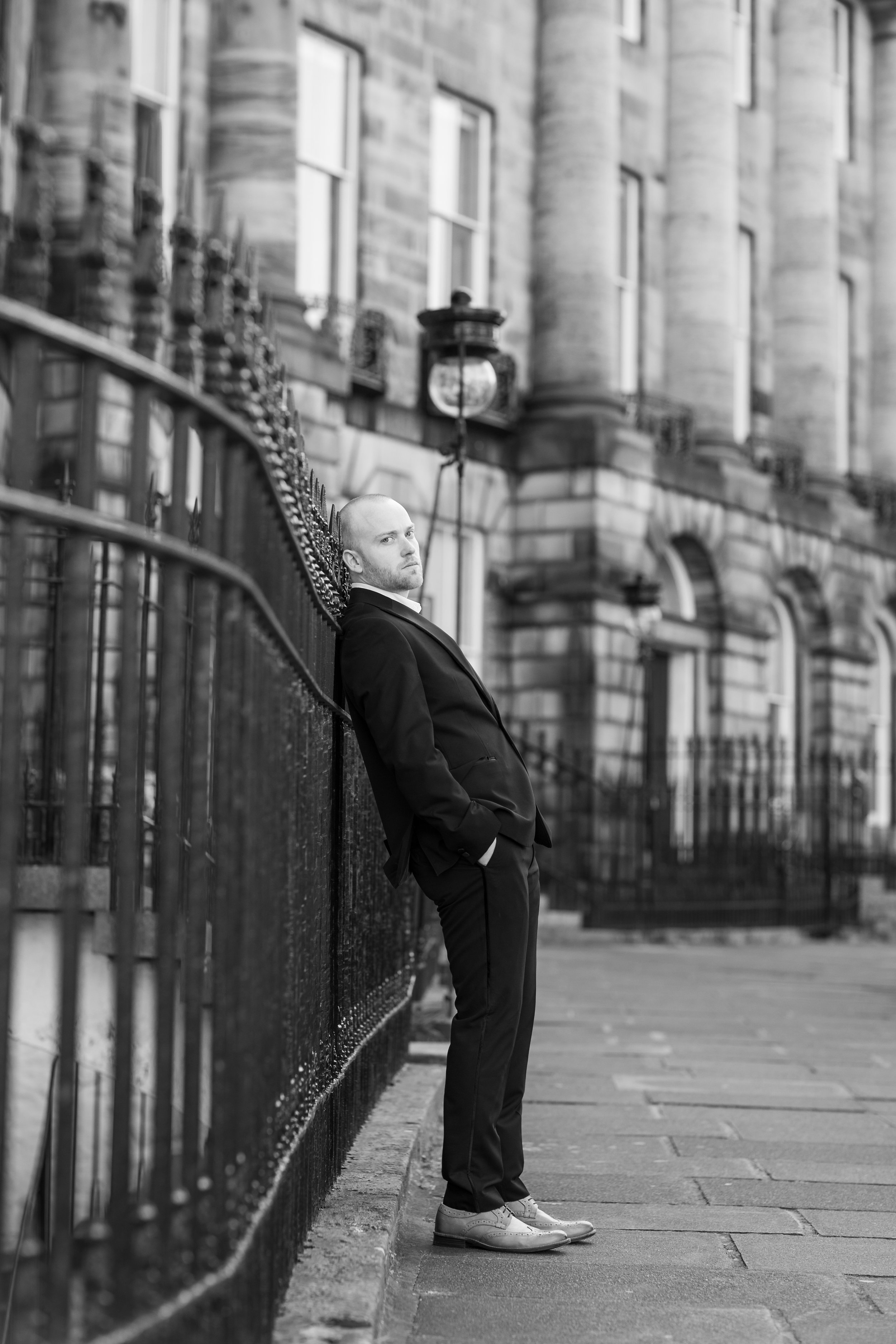 A man in a suit leaning against a black iron fence on a city sidewalk, with a historic stone building in the background.