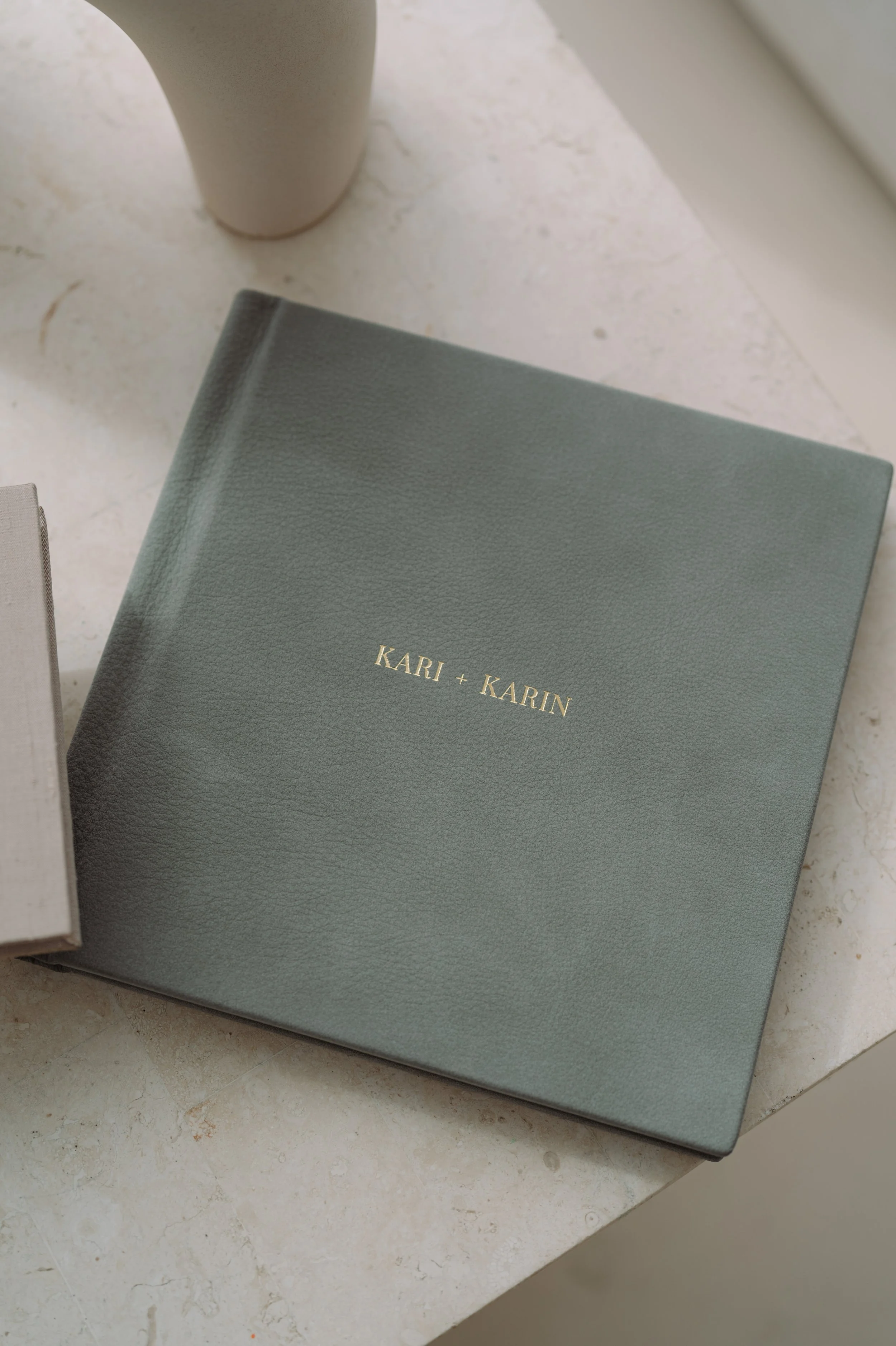 A closed grey book with the title 'KARI + KARIN' embossed in gold on the cover, placed on a light-colored marble surface near a white ceramic vase.