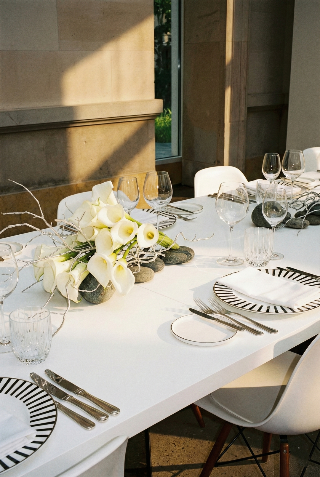 Elegant dining table set with white plates with black striped edges, stainless steel cutlery, clear wine glasses, napkins, and a floral centerpiece of white calla lilies and decorative branches on a white tablecloth, near a large window with sunlight.