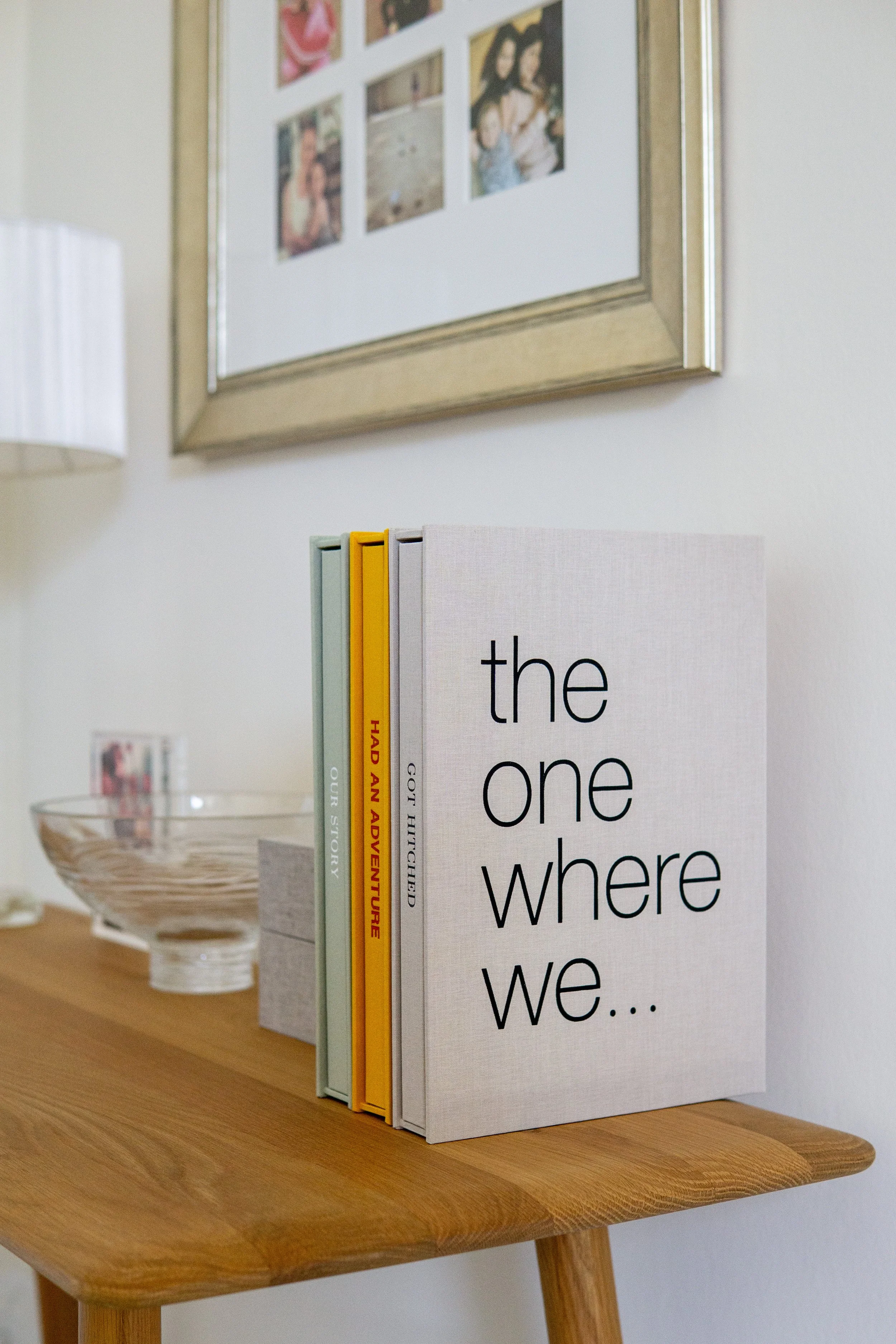 A wooden table with five books, each with different colored covers, standing upright on it. The book titles are partially visible, with the most prominent one reading "the one where we...". In the background, there is a framed photo collage on the wa