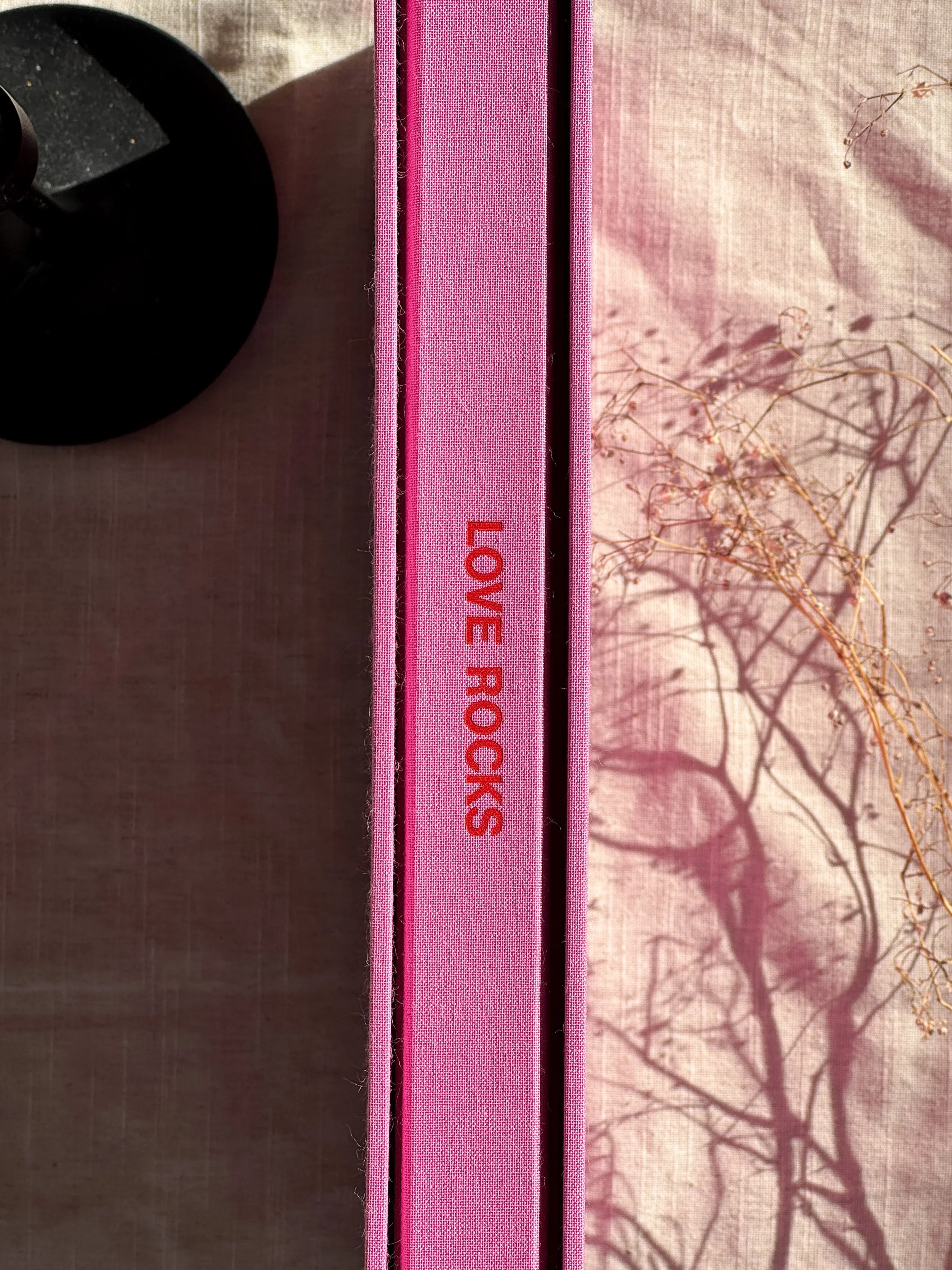 Pink box with the words 'LOVE ROCKS' written in red, placed on a table with a black object and a pink cloth with a shadow of a tree.