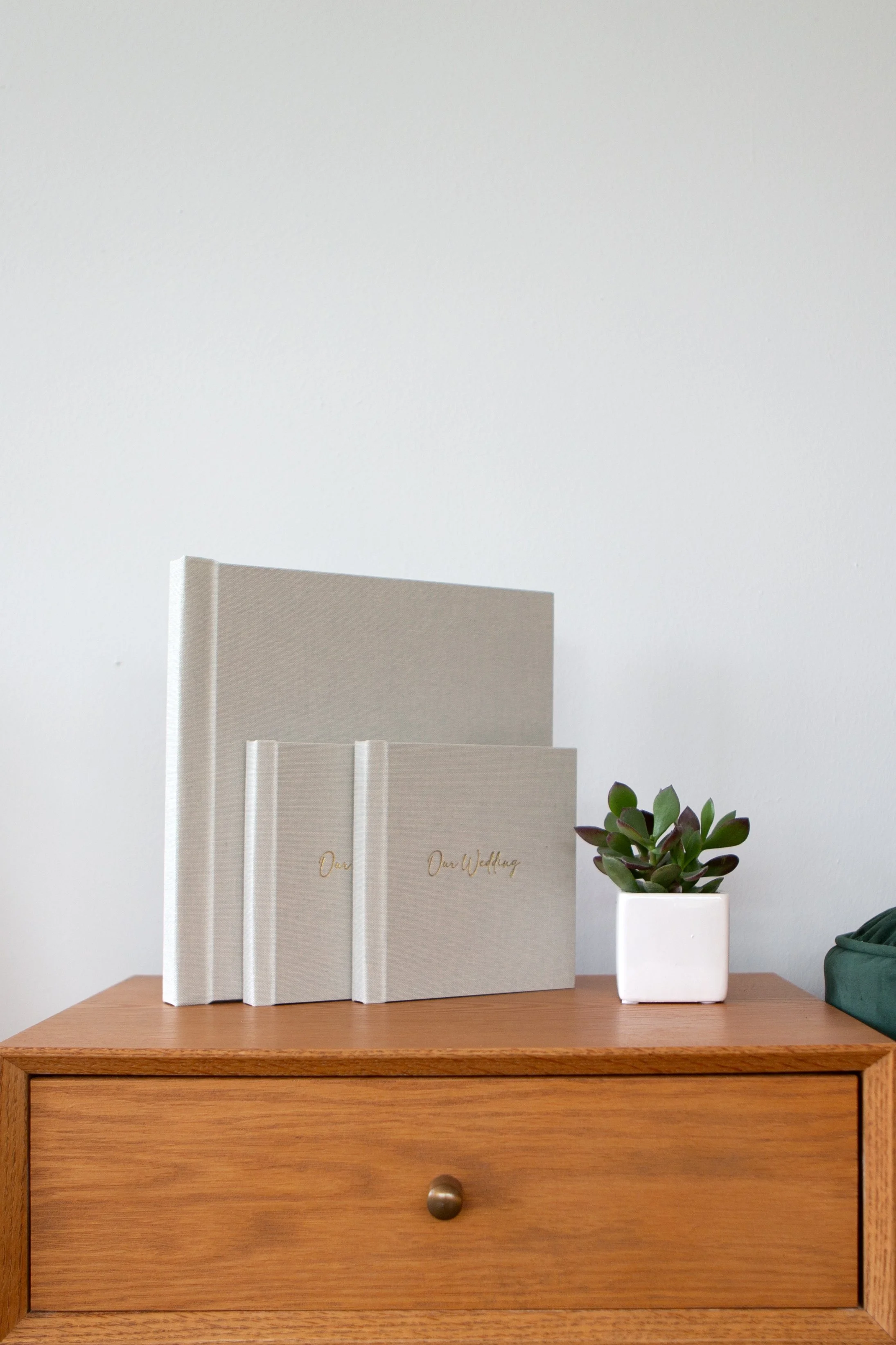 A wooden nightstand holds three beige wedding albums and a potted succulent plant against a white wall.