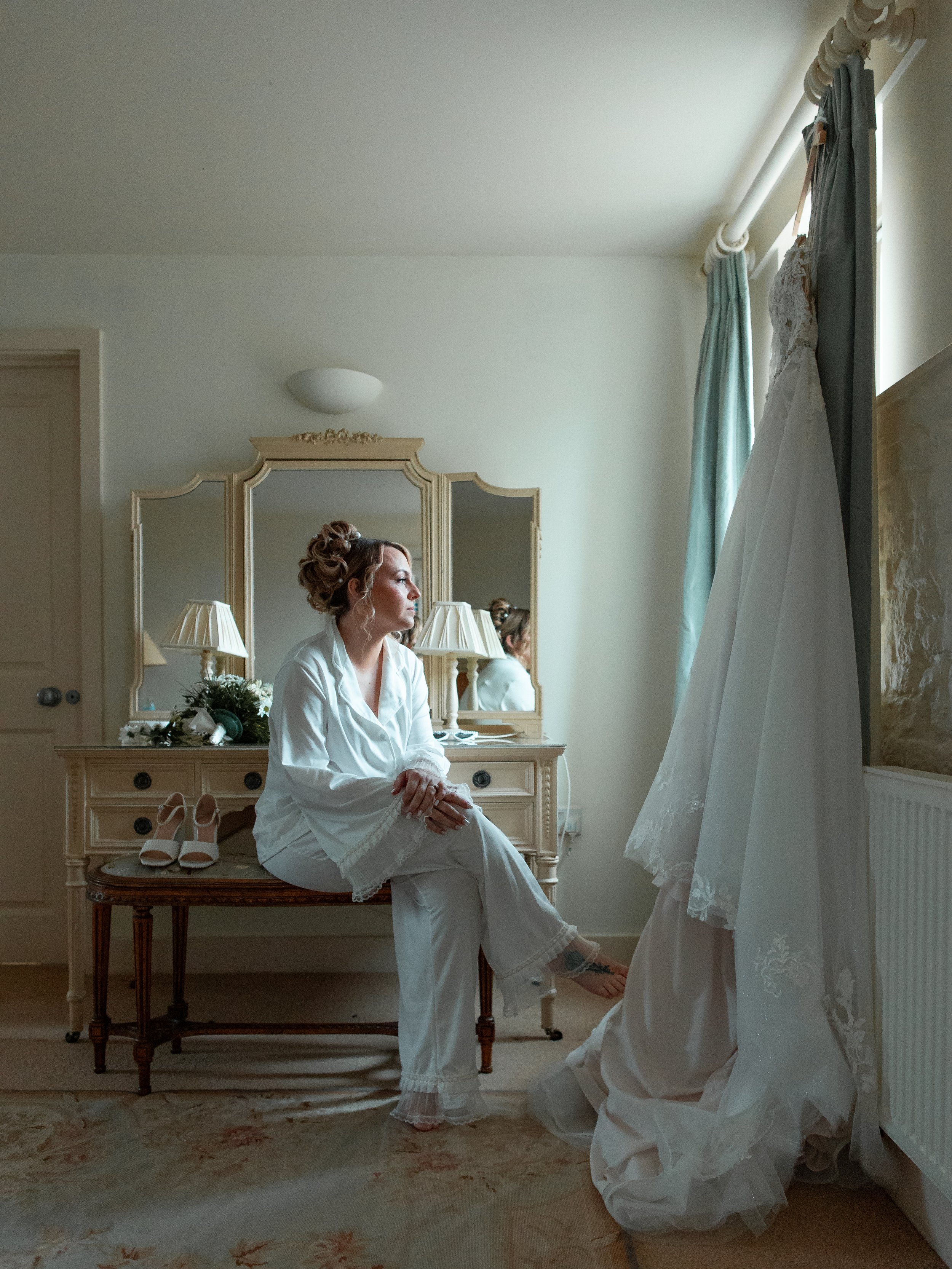 A woman in white pajamas sitting on a vanity bench in a bedroom, looking thoughtfully out the window. A wedding dress is hanging on the curtain rod, and a mirror reflects her profile.