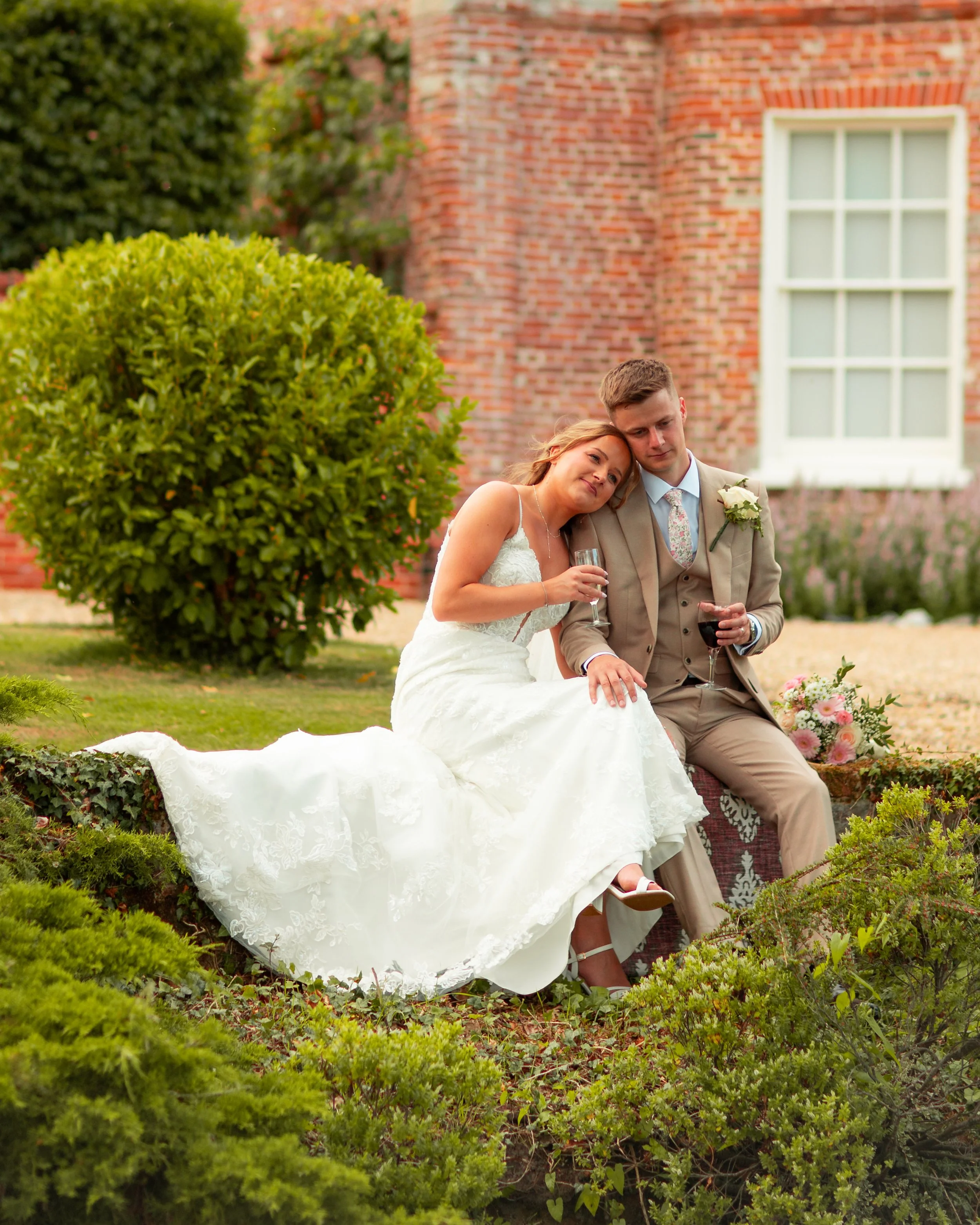 A bride and groom sitting outside on a brick ledge, holding drinks, with a bouquet beside them. They are dressed in wedding attire, appearing emotional and close to each other. The background includes a red brick building, a window, and lush greenery.