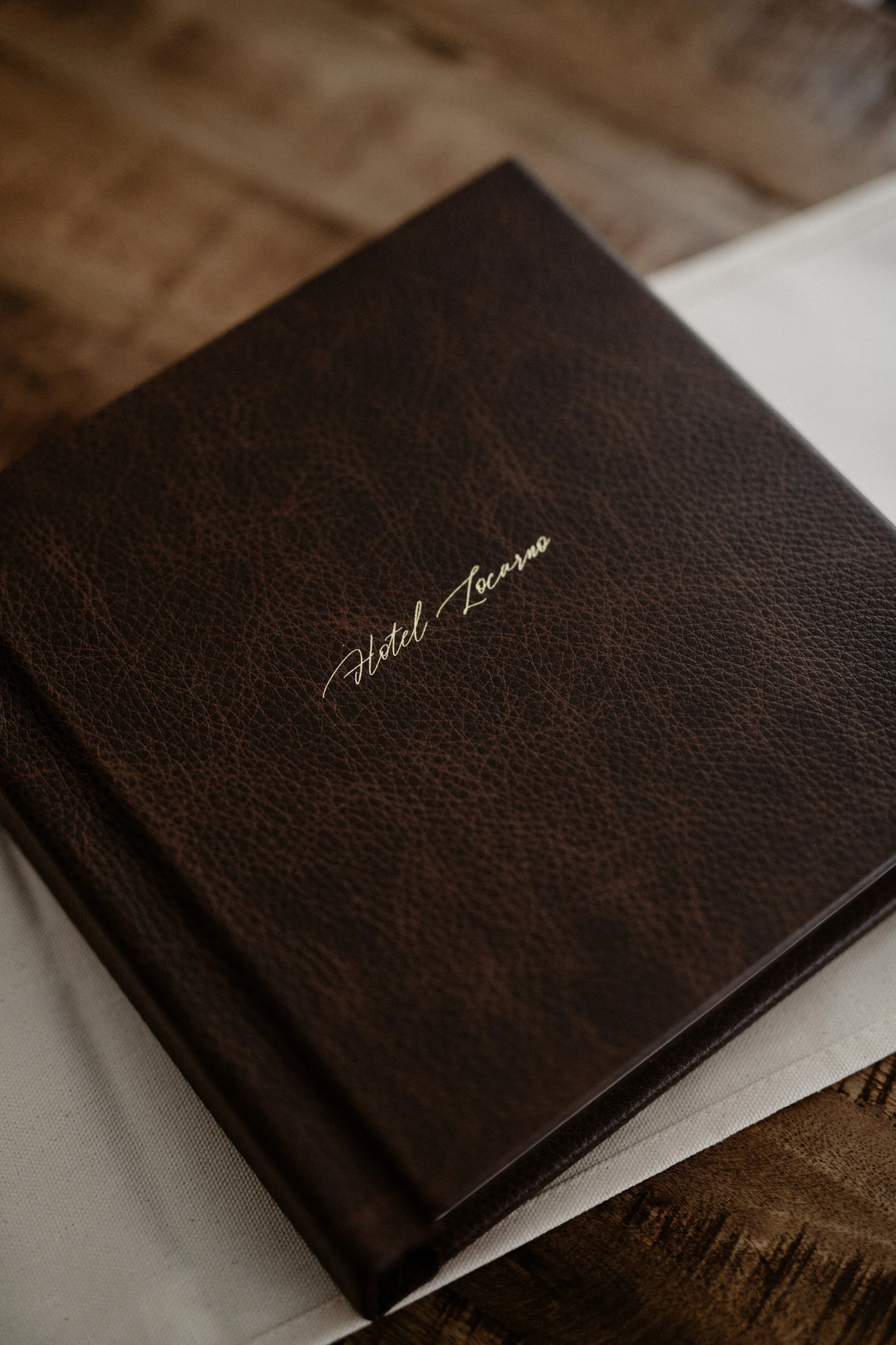 Close-up of a brown leather-bound menu or notebook with 'Hotel Locarno' written in cursive gold lettering on the cover, placed on a white cloth on a wooden table.