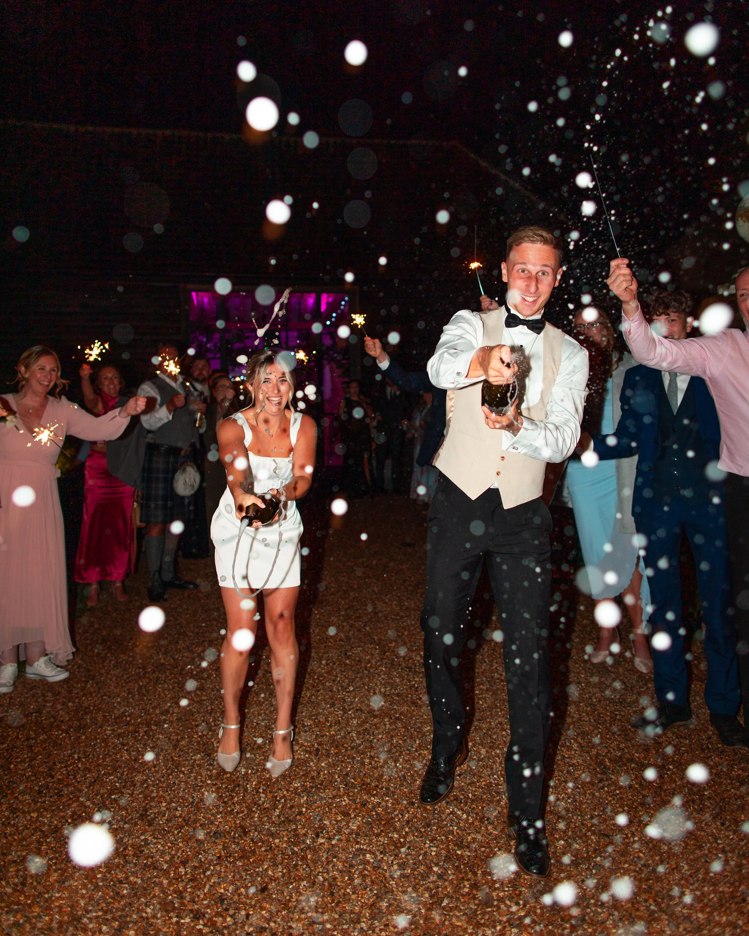 People celebrating a wedding with sparklers and bubbles during the night. The bride and groom are at the center, smiling and holding sparklers, surrounded by guests.