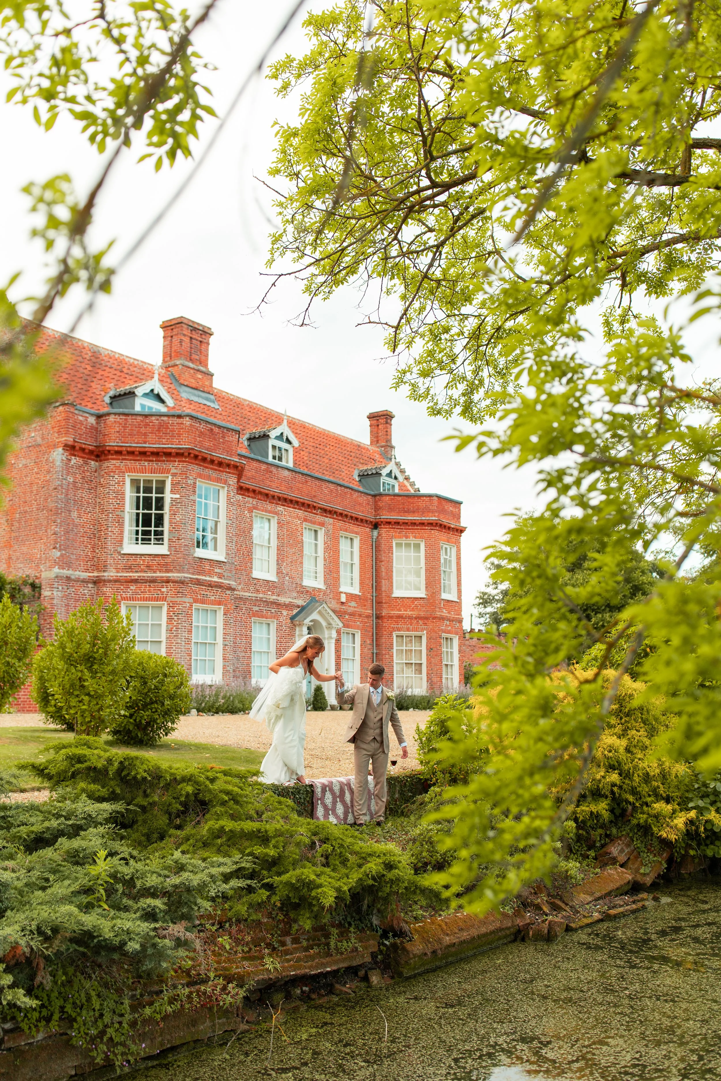 A newlywed couple walking on a garden lawn at Elms Barn, beside a pond, with a large red brick mansion in the background, framed by green trees.