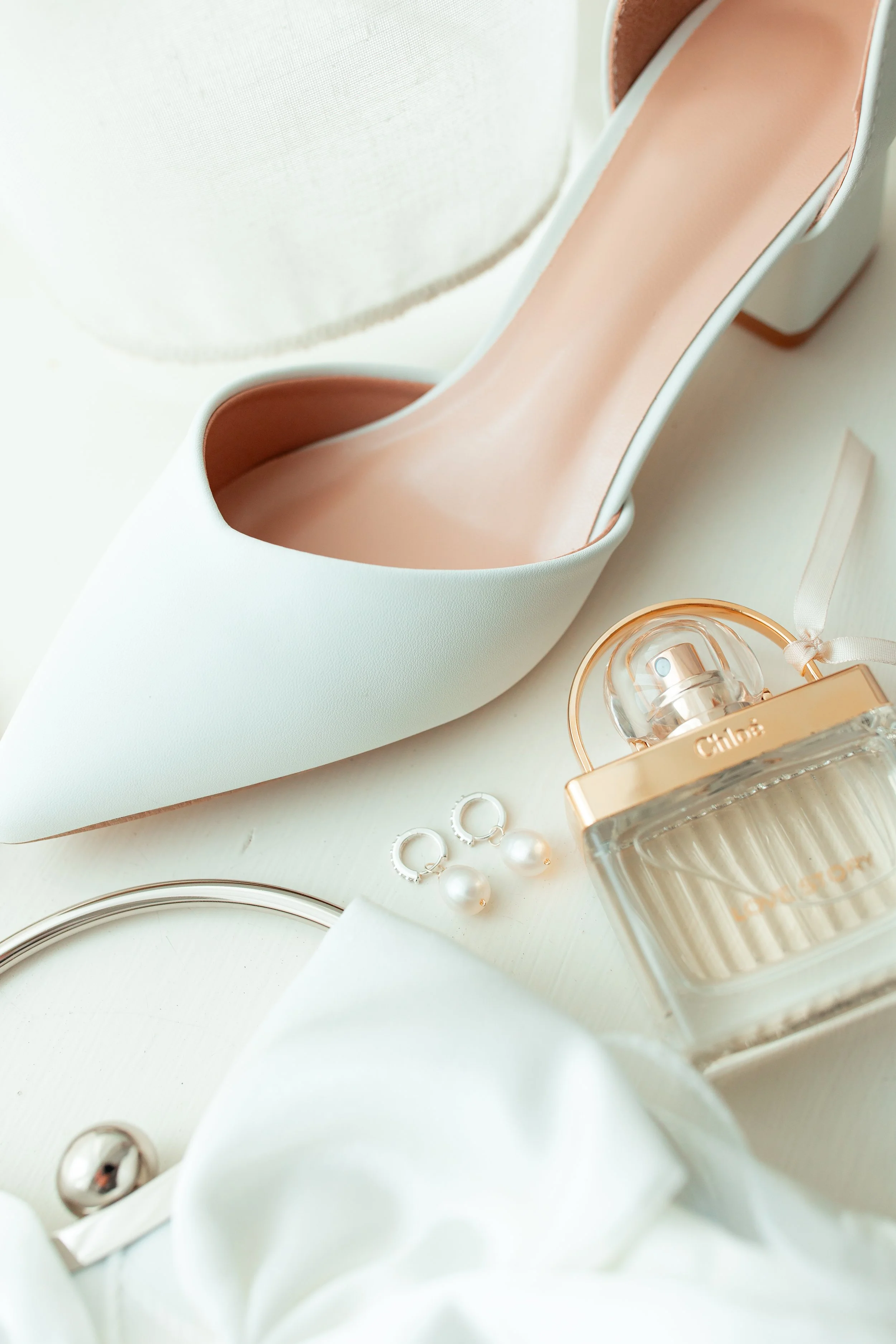 A white high heel shoe, a bottle of Chloe perfume, pearl earrings, and a white clutch purse arranged on a white surface.
