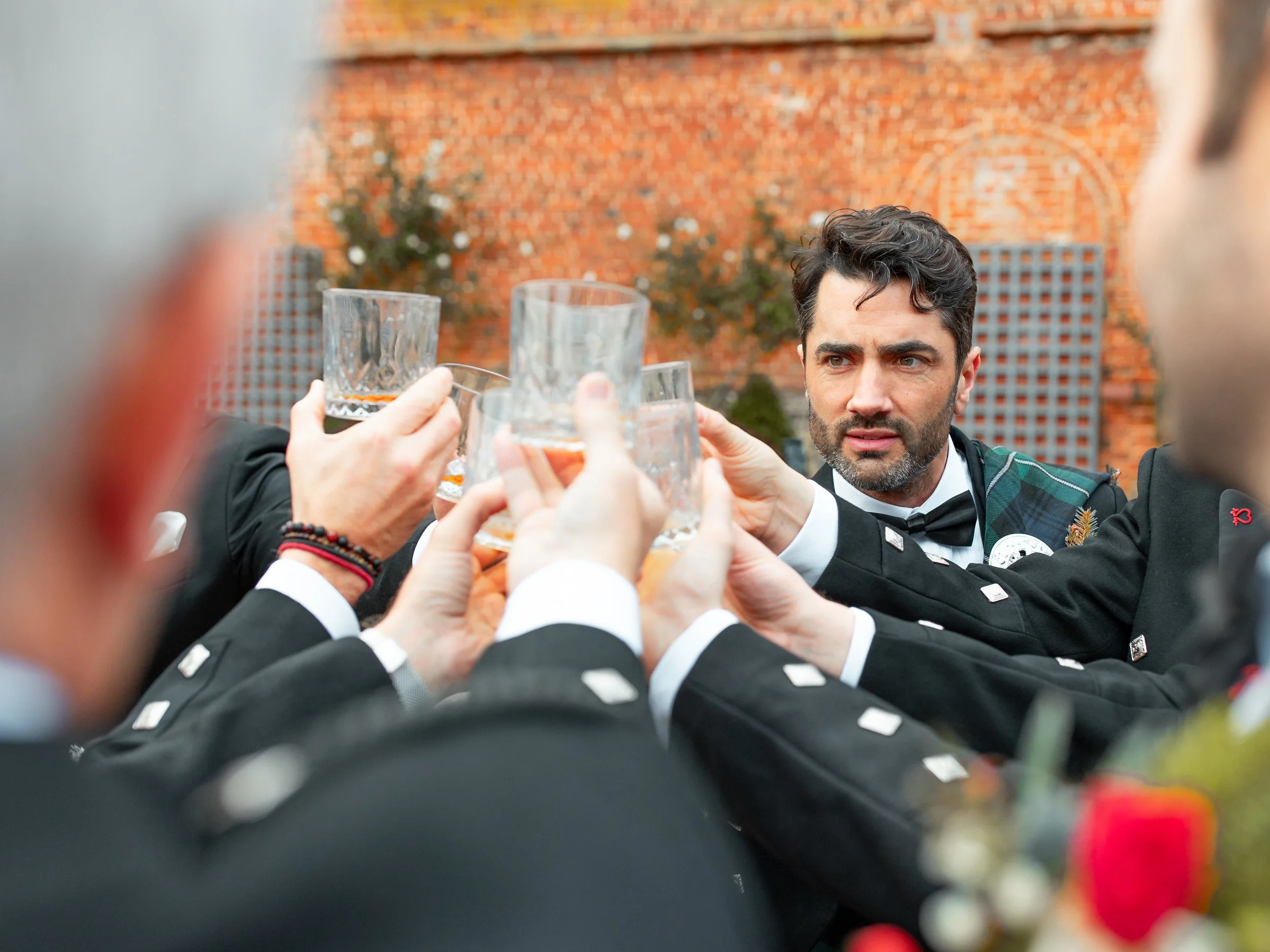 A man in a tuxedo and tartan sash looks serious as he toasts with others in uniform at an outdoor gathering, holding glasses of drink against a brick wall background.