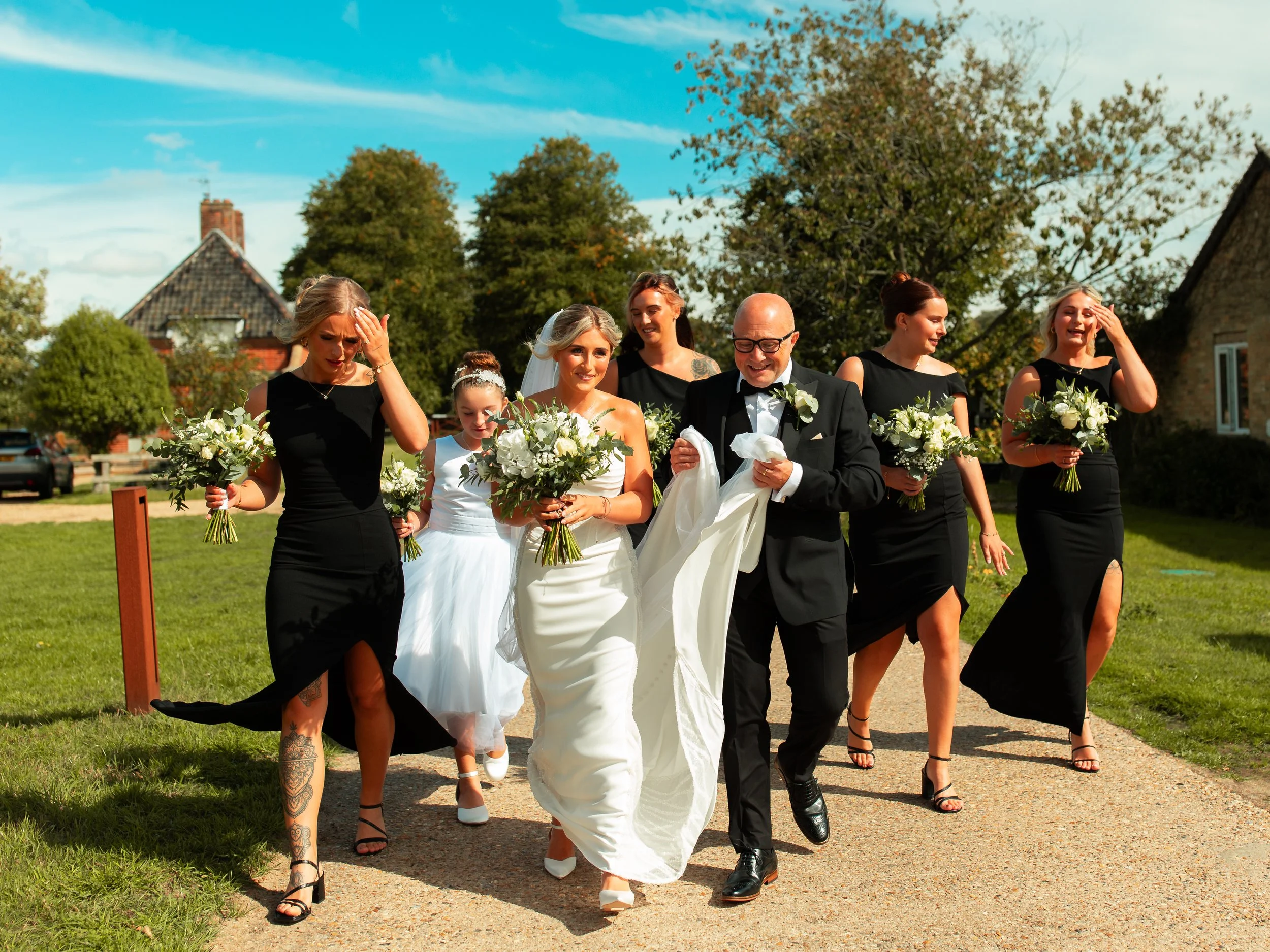 A bride in a white wedding dress walking outdoors with her wedding party, including bridesmaids in black dresses holding bouquets, and a groom in a tuxedo, on a sunny day with trees and houses in the background.