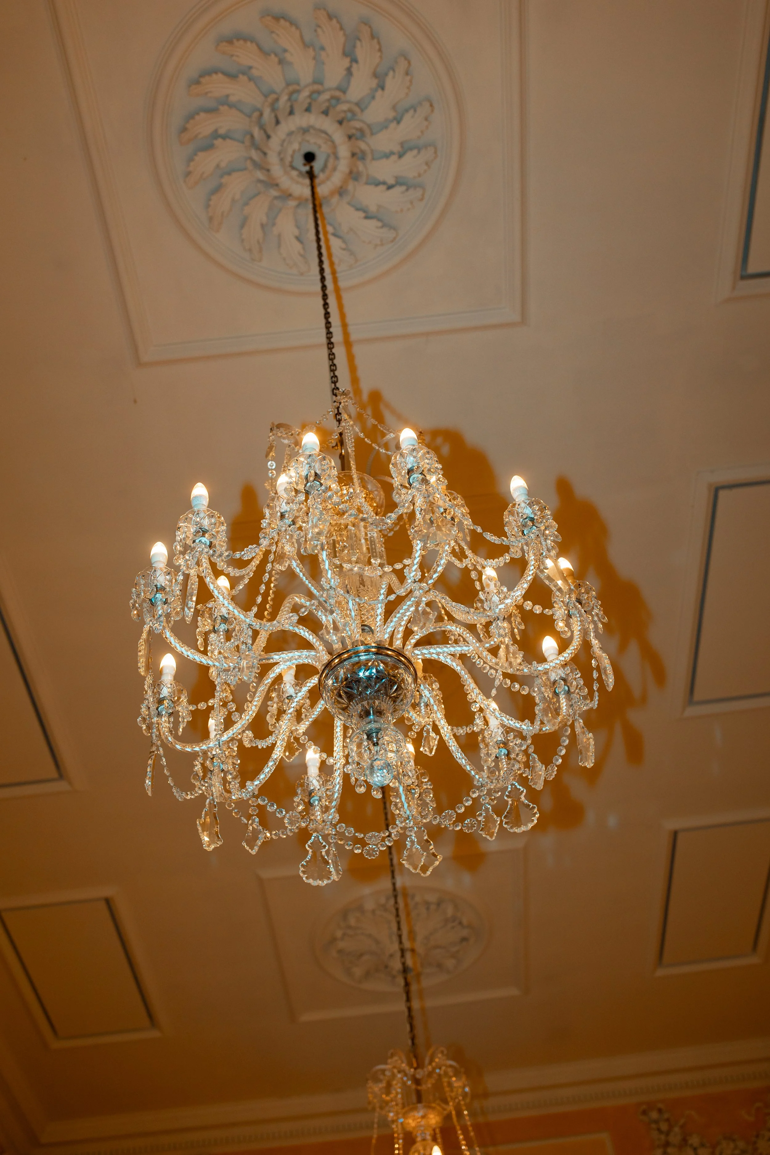 A large, ornate crystal chandelier hanging from a decorated ceiling, illuminated with multiple small bulbs, casting shadows on the ceiling.