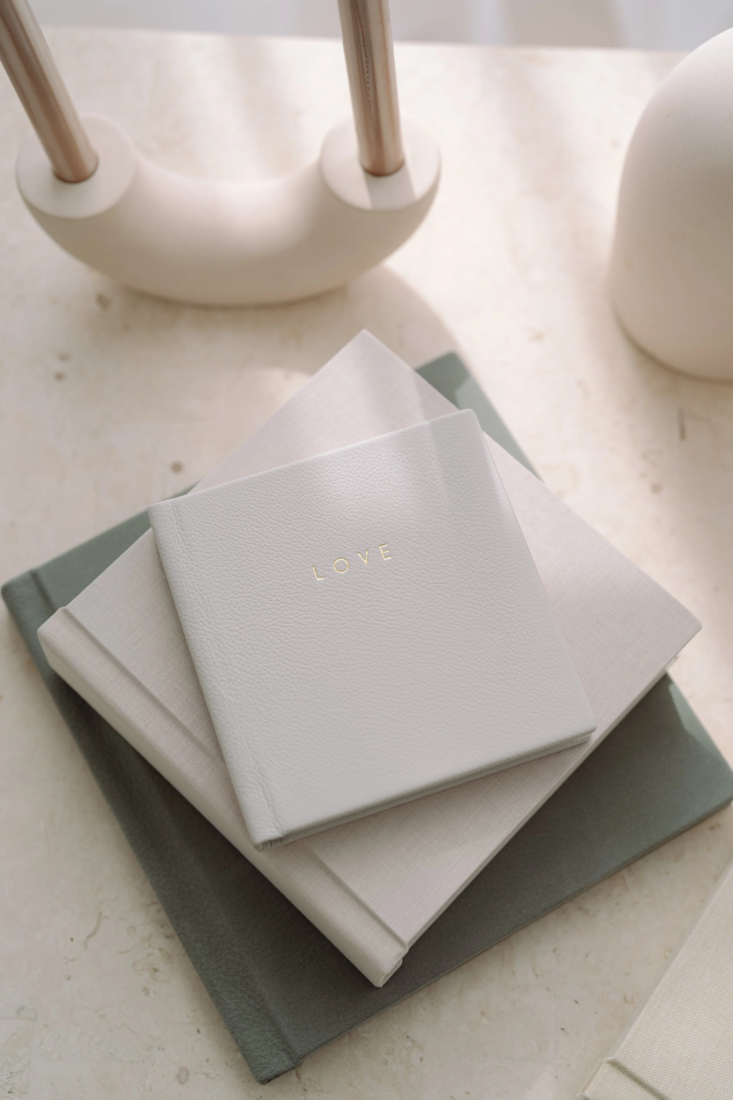 A stack of three fabric-covered books or boxes in shades of beige and gray, with a white textured napkin or cloth on top, featuring the word 'LOVE' in gold lettering. The background includes a white table surface and decorative objects, including a white ceramic holder with two metal rods and a white rounded container.