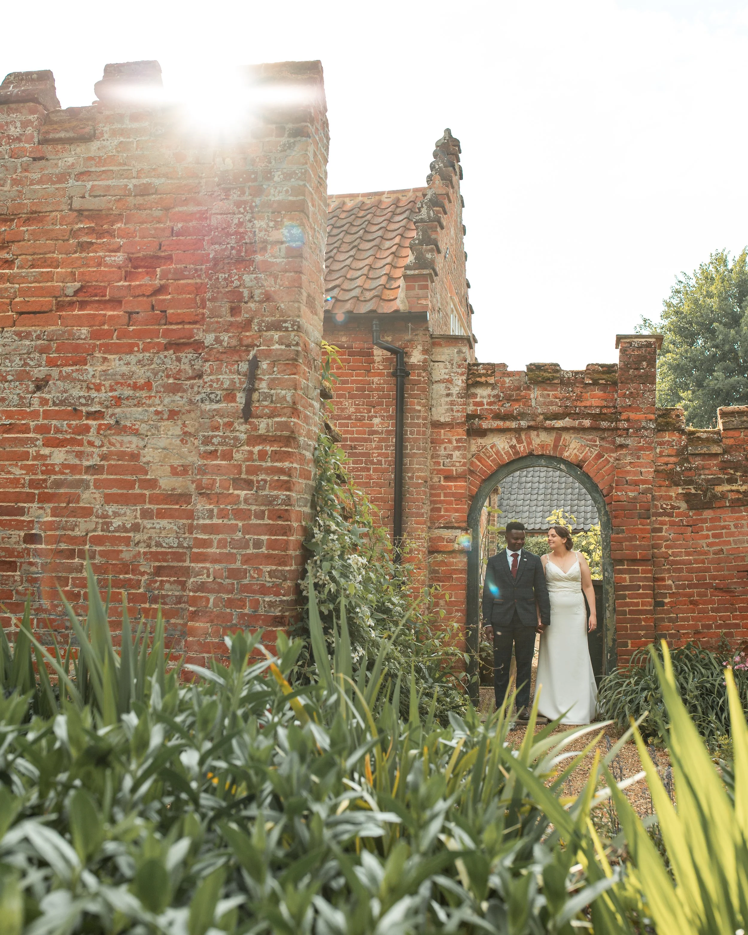 A bride and groom walk hand-in-hand through a garden archway in front of an old brick building on a sunny day.