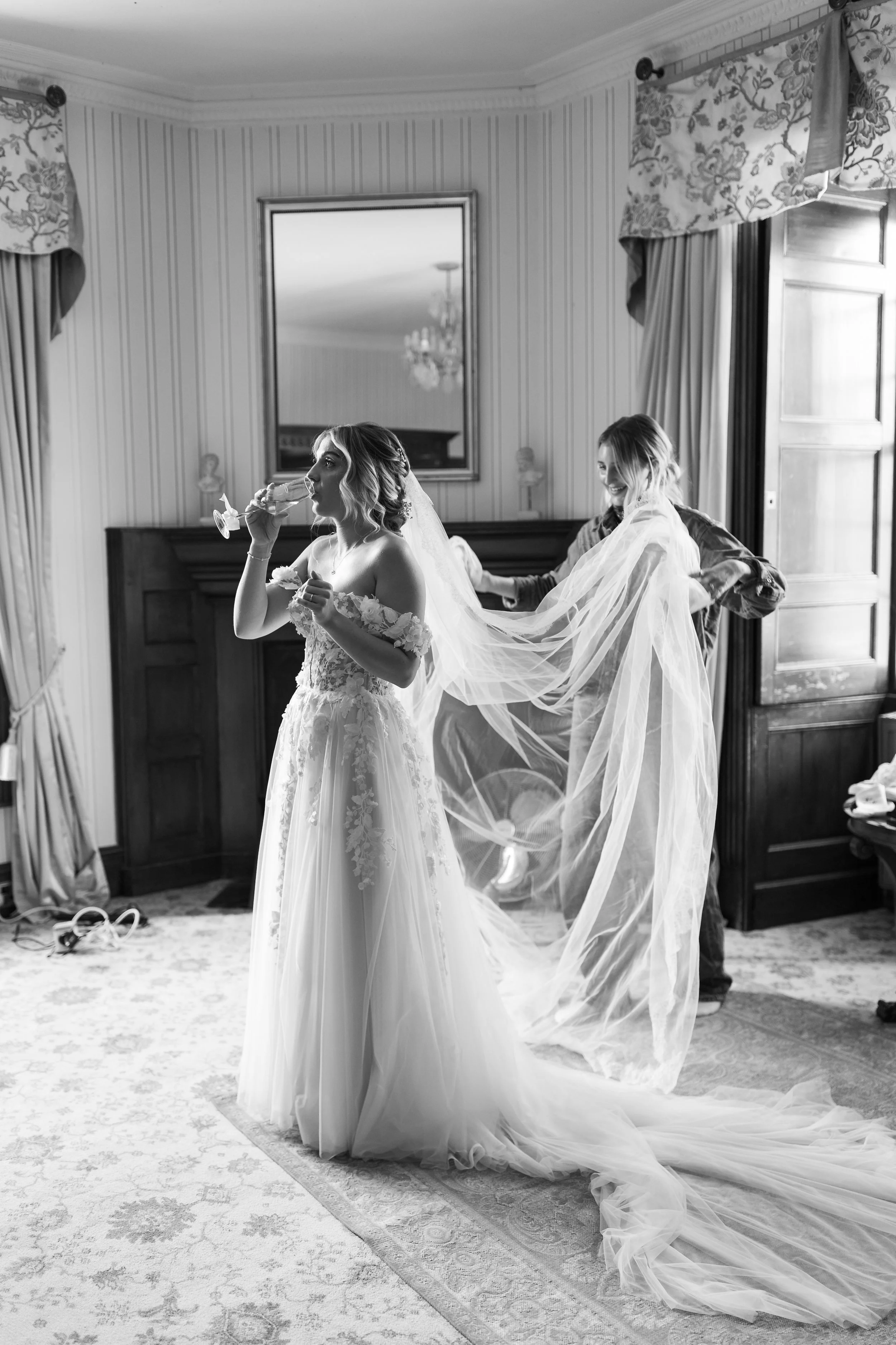 Black and white photo of a bride in a wedding gown, playing with a glass of champagne while a woman behind her helps with her veil in a decorated room.