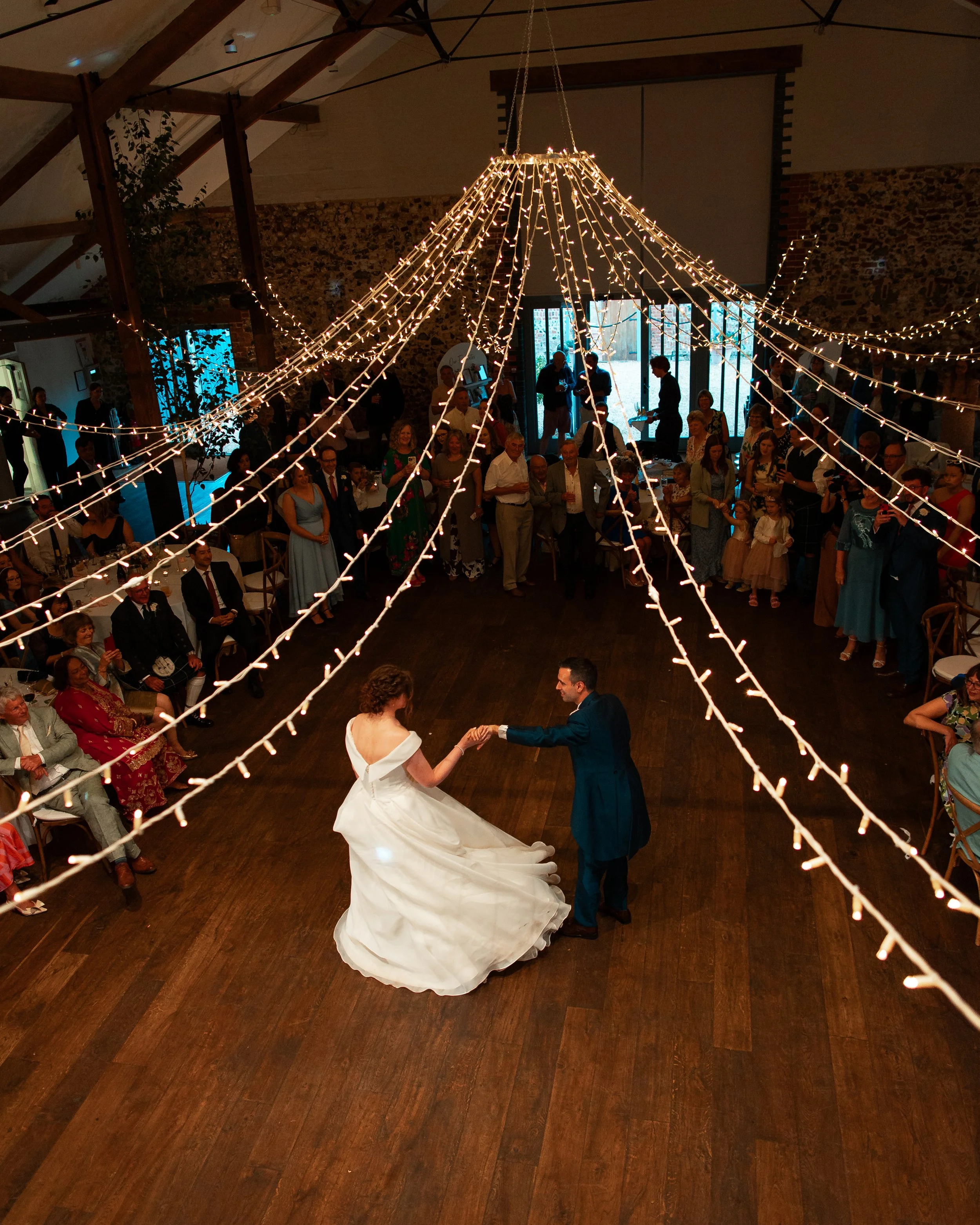A bride and groom dancing in the center of a wedding reception hall with guests watching, illuminated by string lights hanging from the ceiling.