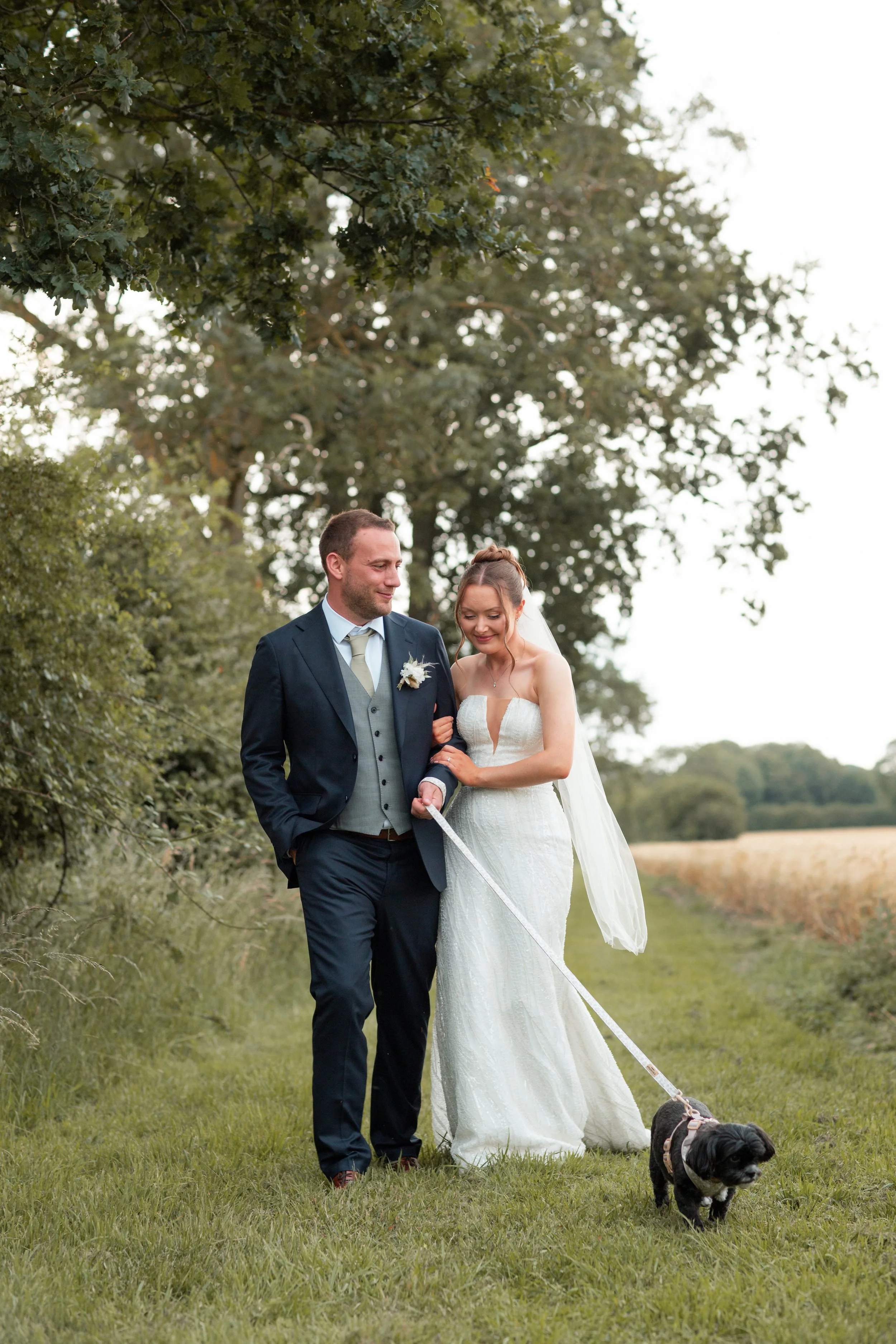 A bride and groom walk outdoors, holding hands. The bride is in a white wedding dress, and the groom is in a dark suit. They are accompanied by a cute black and white dog on a leash in a grassy area with trees and open land in the background.