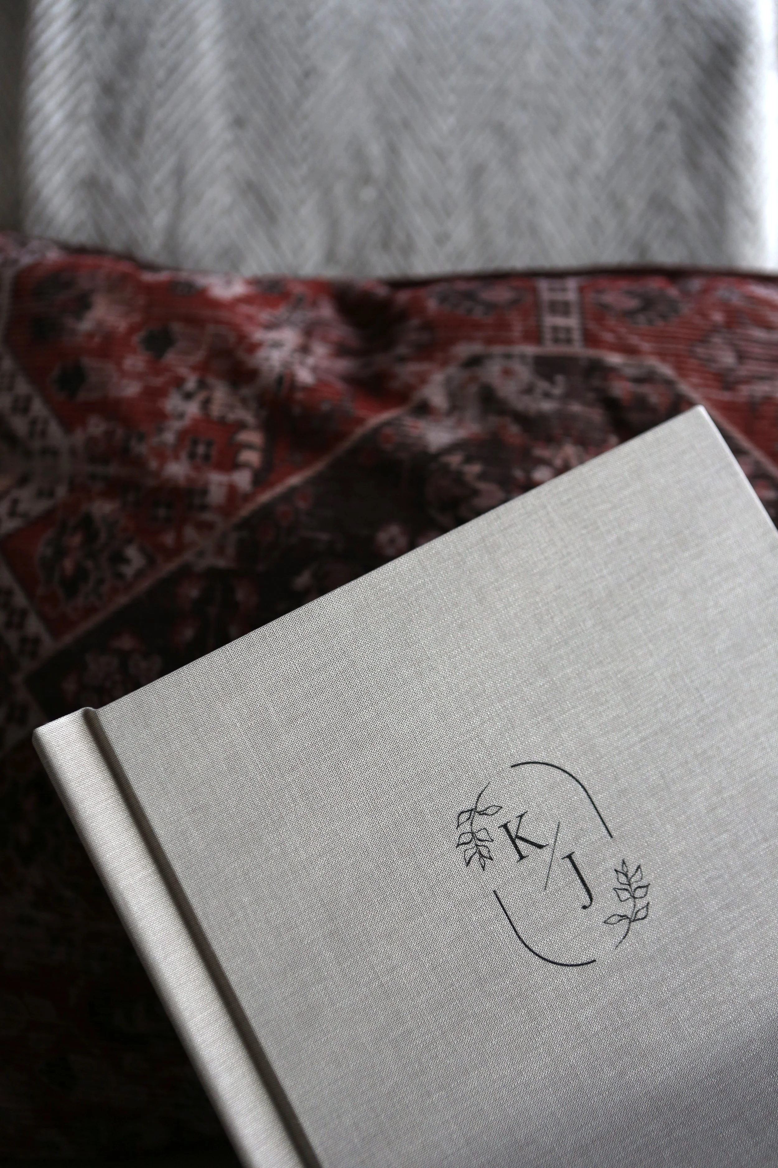 A light-colored, textured notebook with initials 'K' and 'J' surrounded by a decorative leaf design on the cover, held over a patterned fabric with red, black, and white colors.