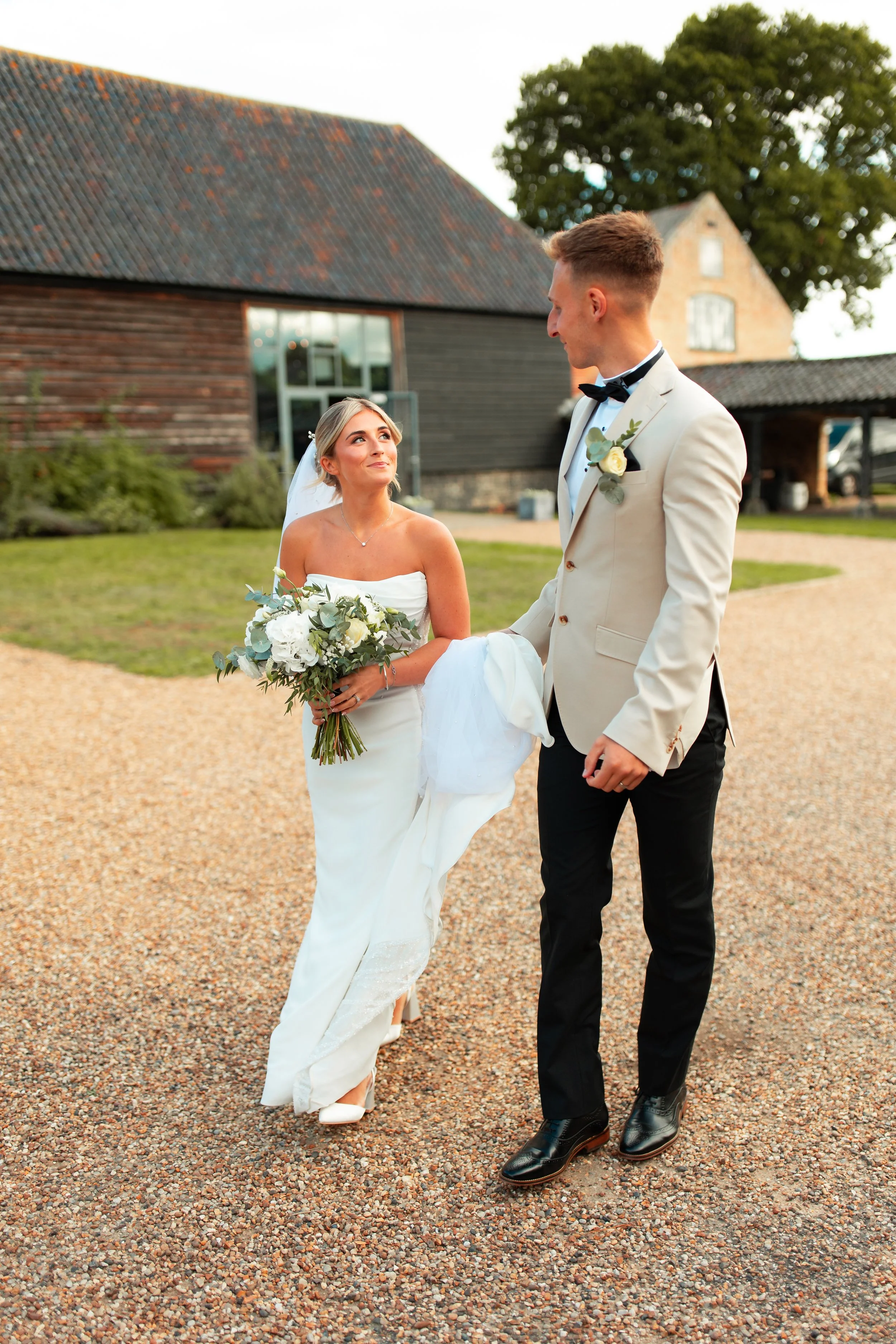 Bride in a white wedding dress holding a bouquet looking at groom in a beige tuxedo with a boutonniere outdoors.