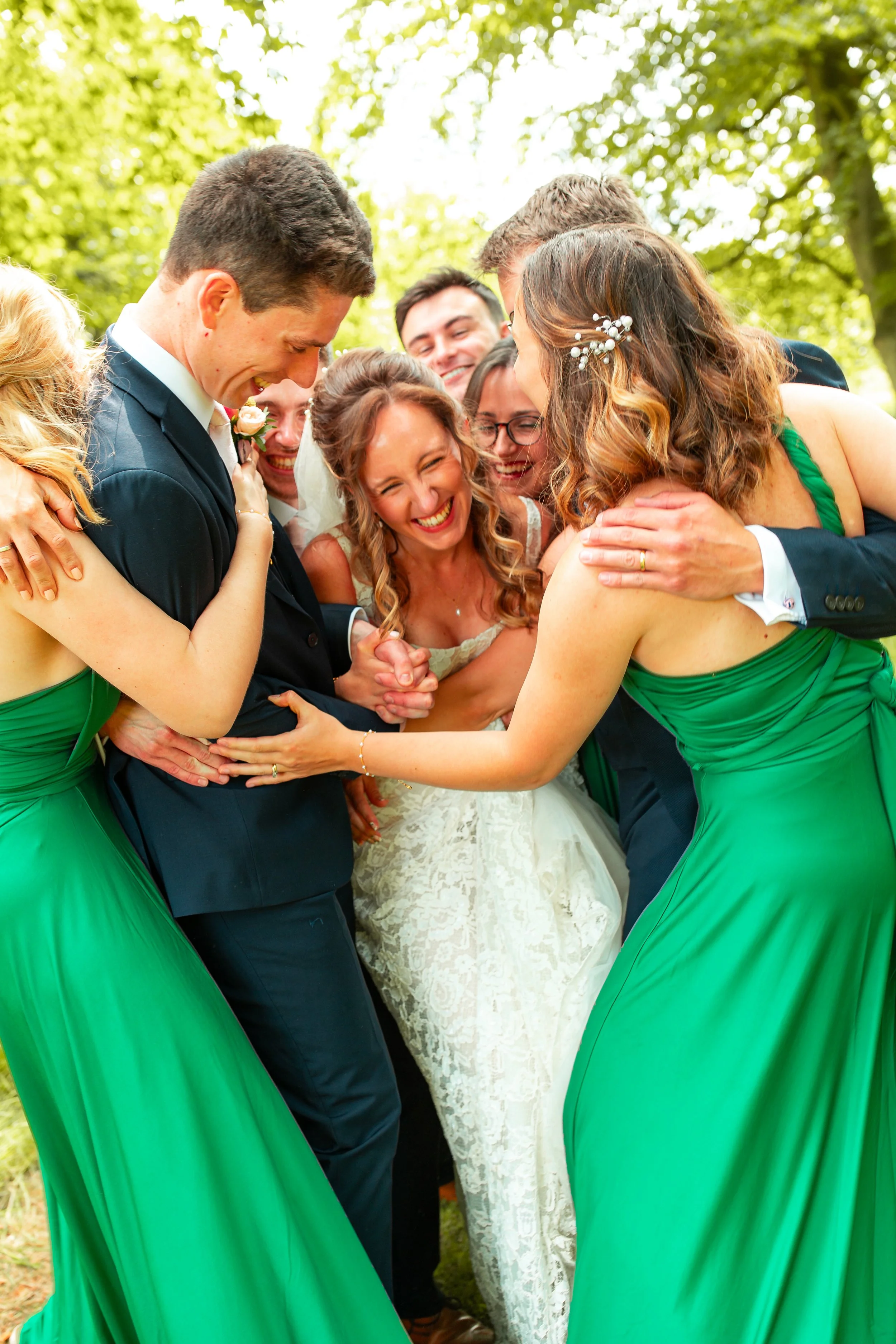 Group of wedding friends and family laughing and hugging outdoors, bride in a white dress, groom in a black suit, surrounded by women in green dresses and men in dark suits, under green trees.