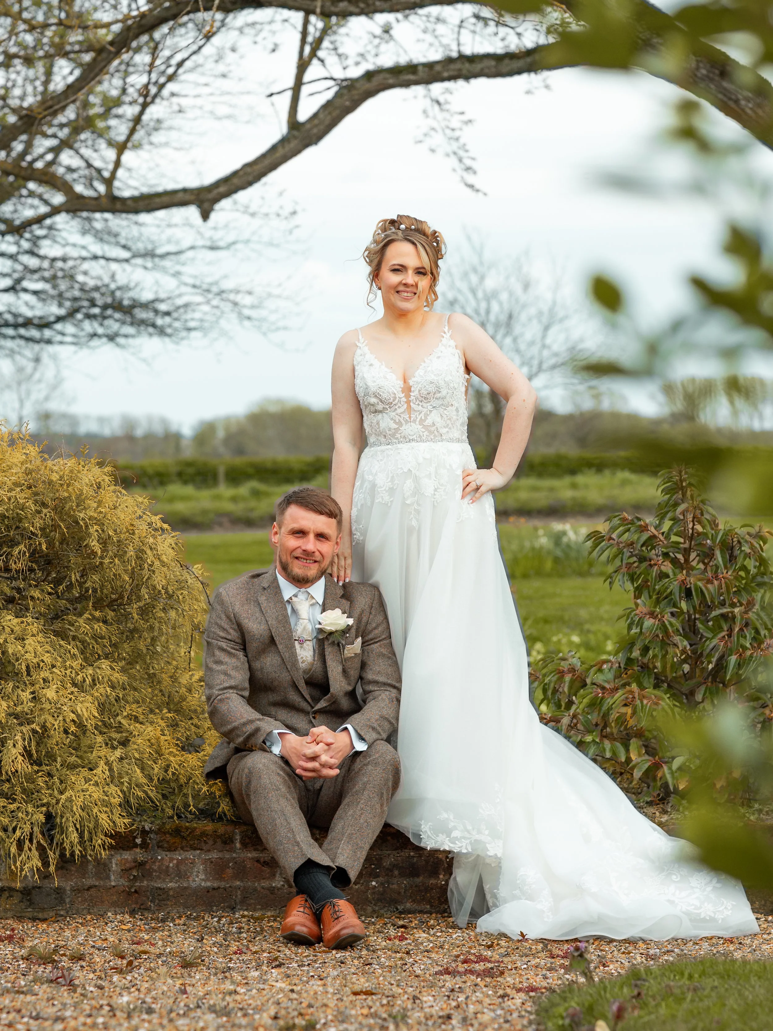 A bride in a white lace wedding dress standing next to a groom in a brown tweed suit, sitting on a brick wall outdoors during daytime.
