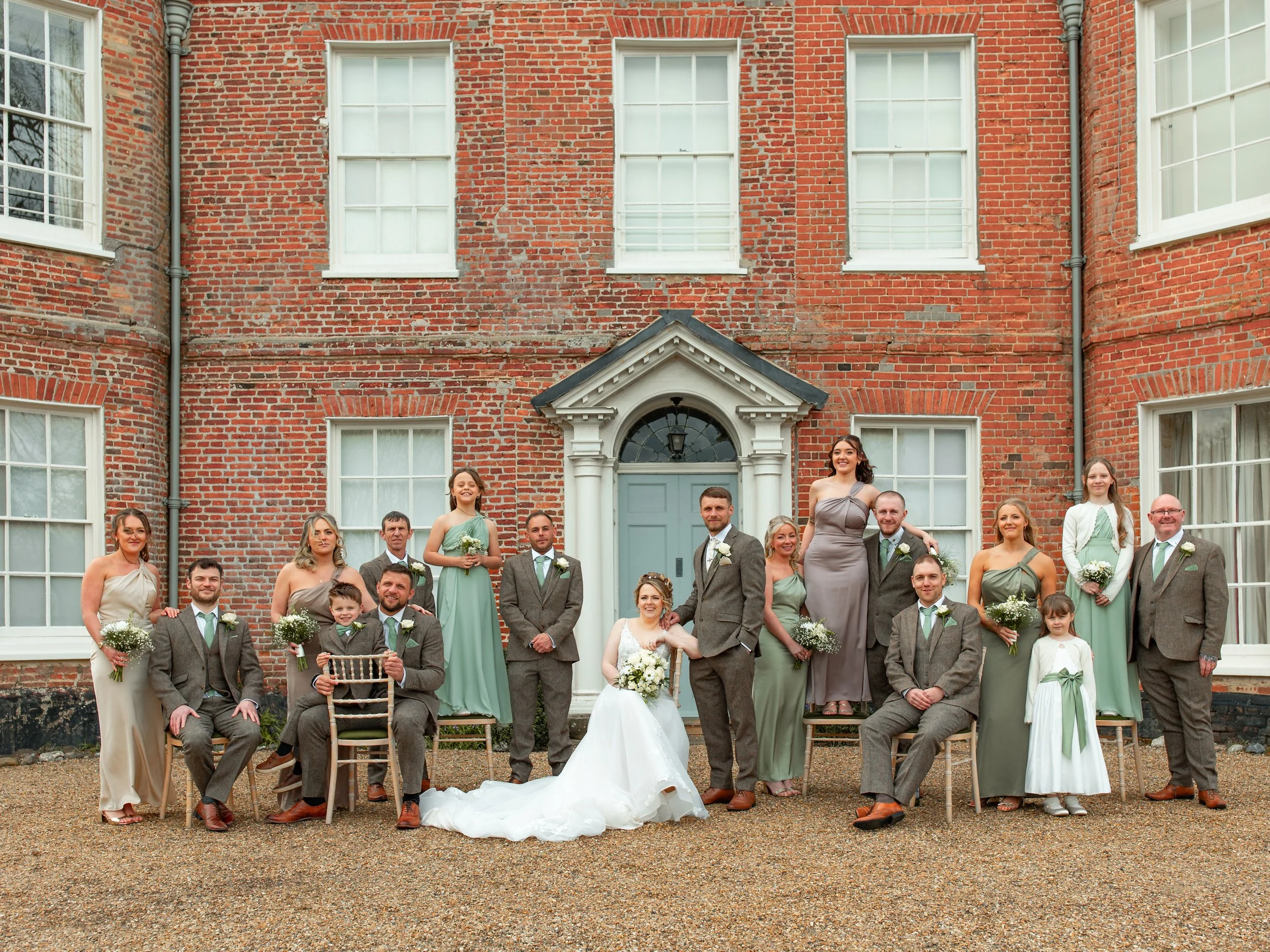 A large wedding party in front of a brick building with multiple windows. The group includes brides, grooms, bridesmaids, groomsmen, children, and family members dressed in formal and semi-formal attire.