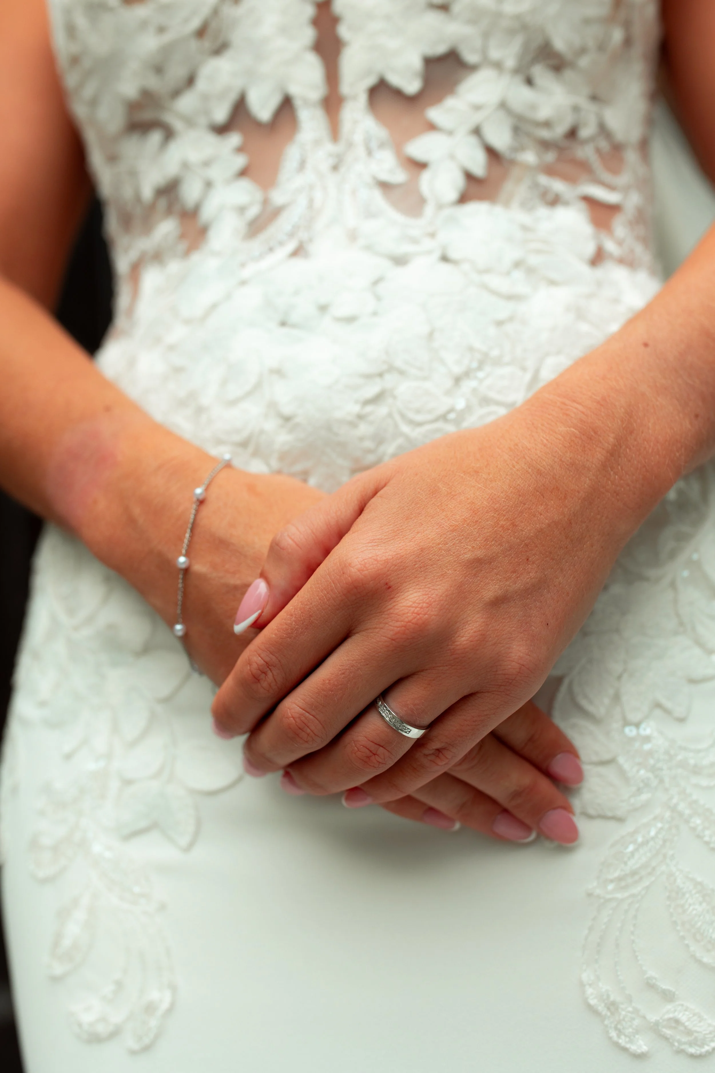 Close-up of a woman's hands with a wedding ring, resting on her lap in a wedding dress with lace details.