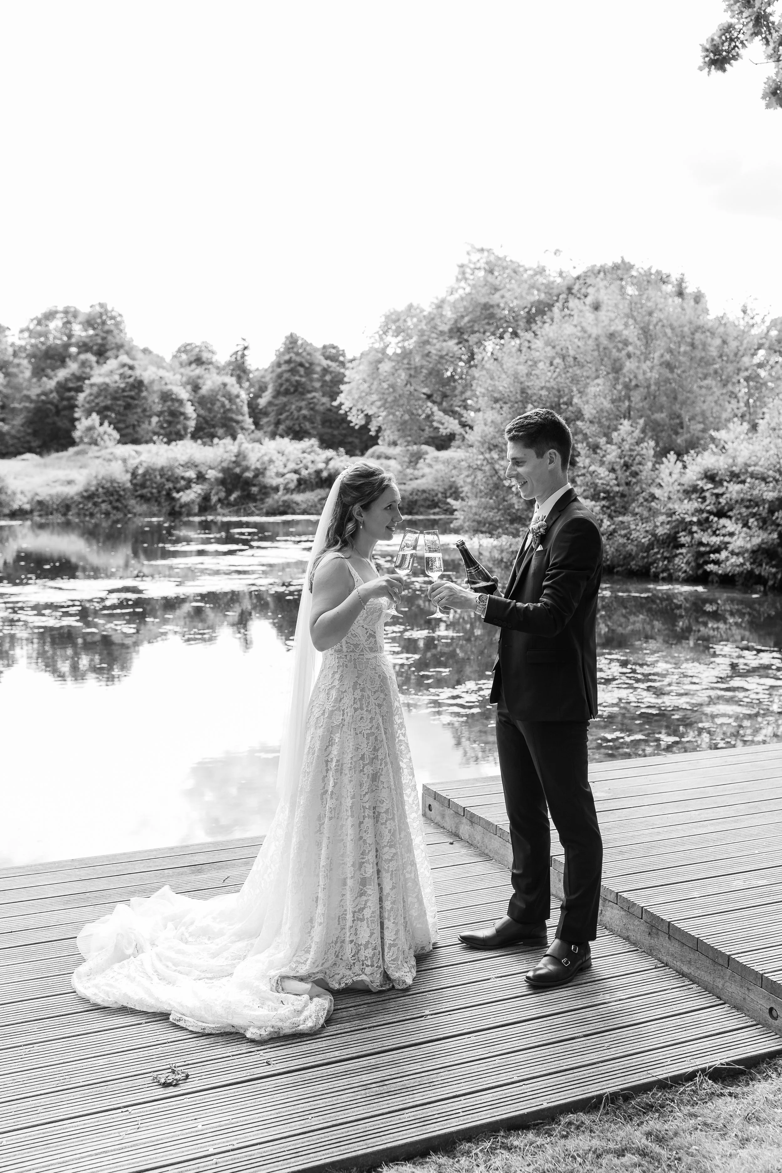 A black-and-white photo of a bride and groom celebrating with drinks on a wooden dock by a lake, with trees in the background.