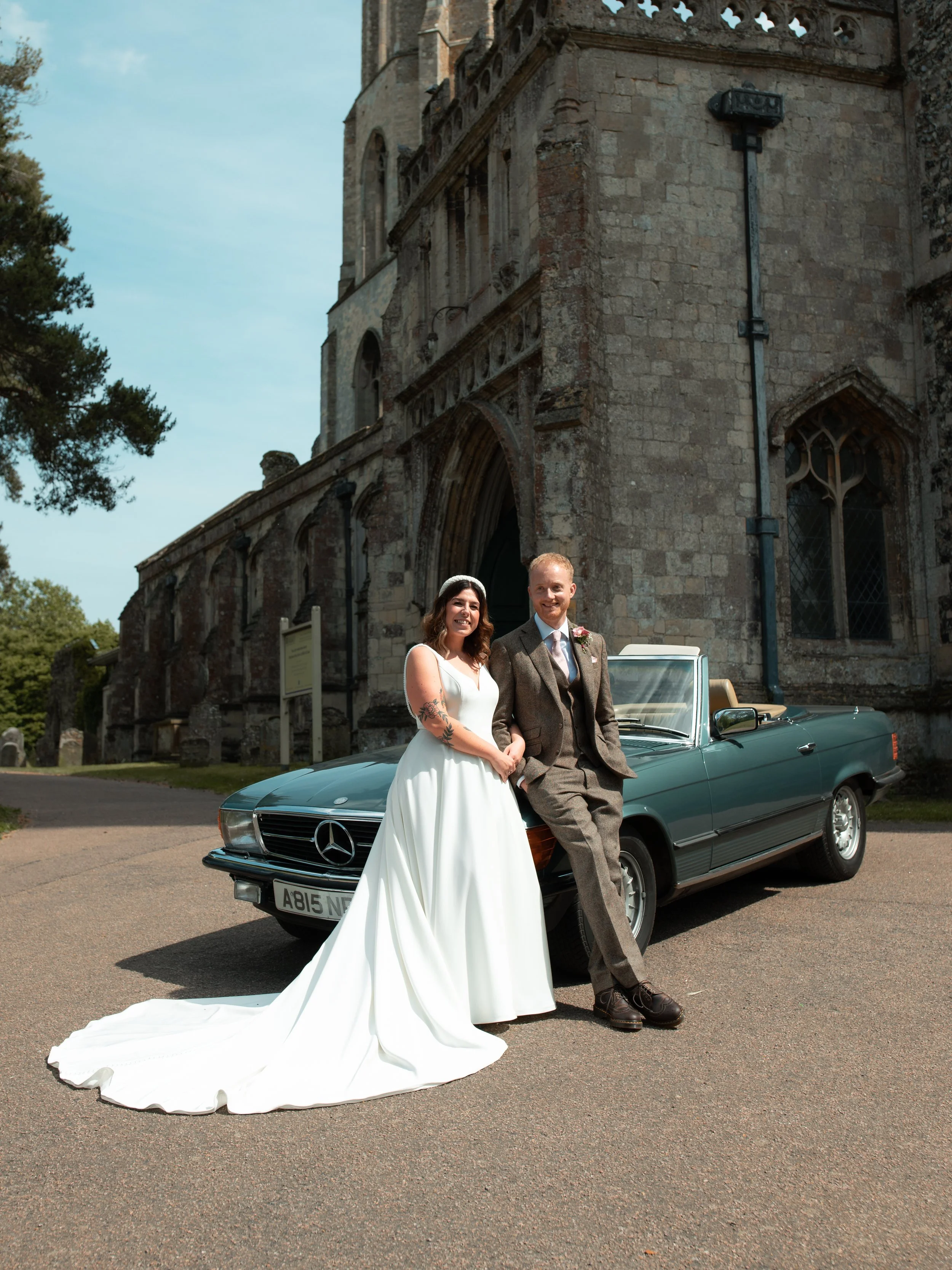 A bride and groom standing in front of a vintage Mercedes-Benz car outside a historic stone church, with the bride in a white wedding dress and the groom in a brown suit, both smiling.