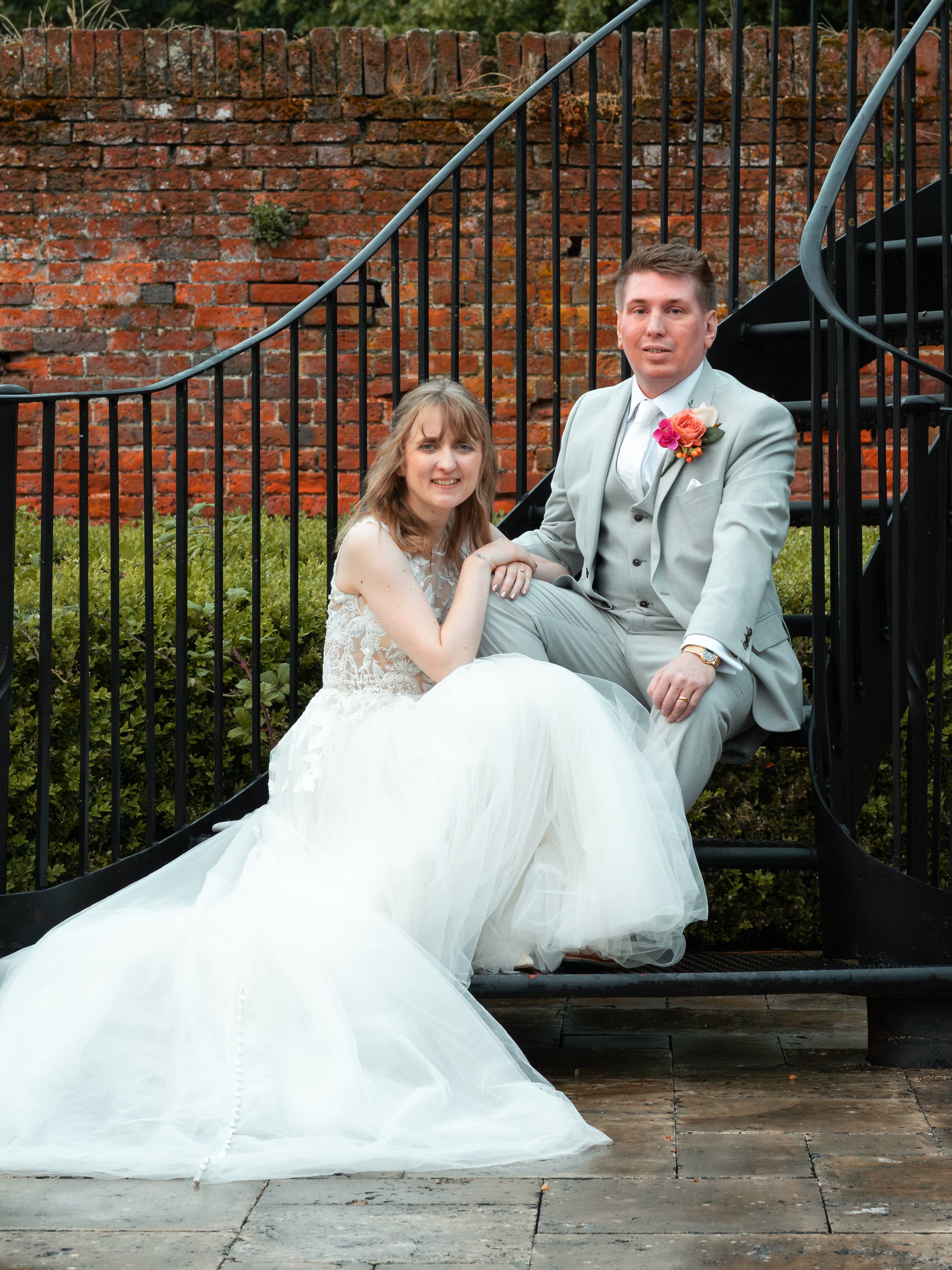 A bride and groom sitting on black metal stairs outdoors, with the bride in a white wedding dress and the groom in a light gray suit with a floral boutonniere. Behind them is a red brick wall and some greenery.
