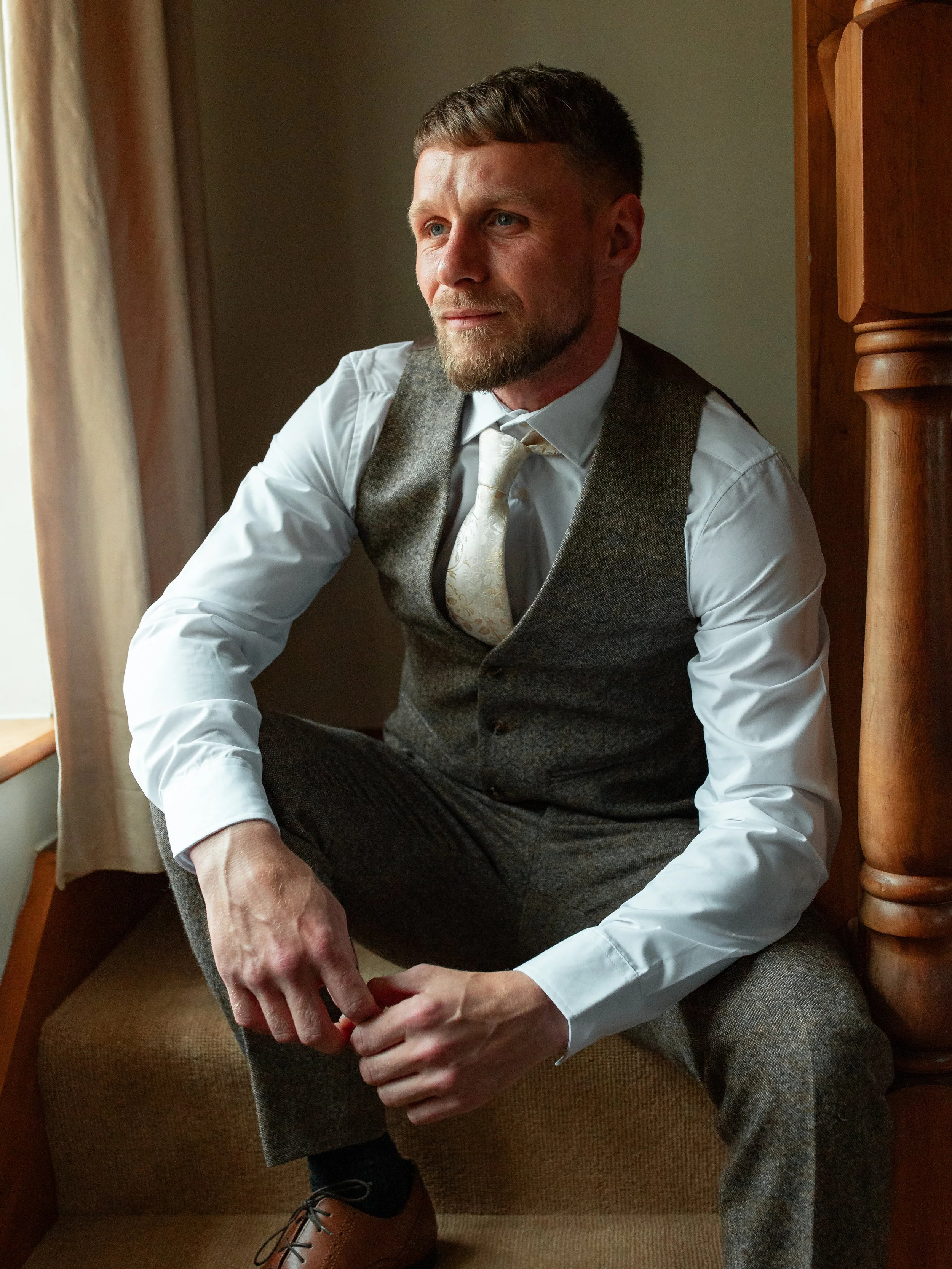 A man in a gray vest and dress pants sits on a window seat, looking thoughtfully out the window, dressed in formal attire including a white shirt, tie, and brown shoes.