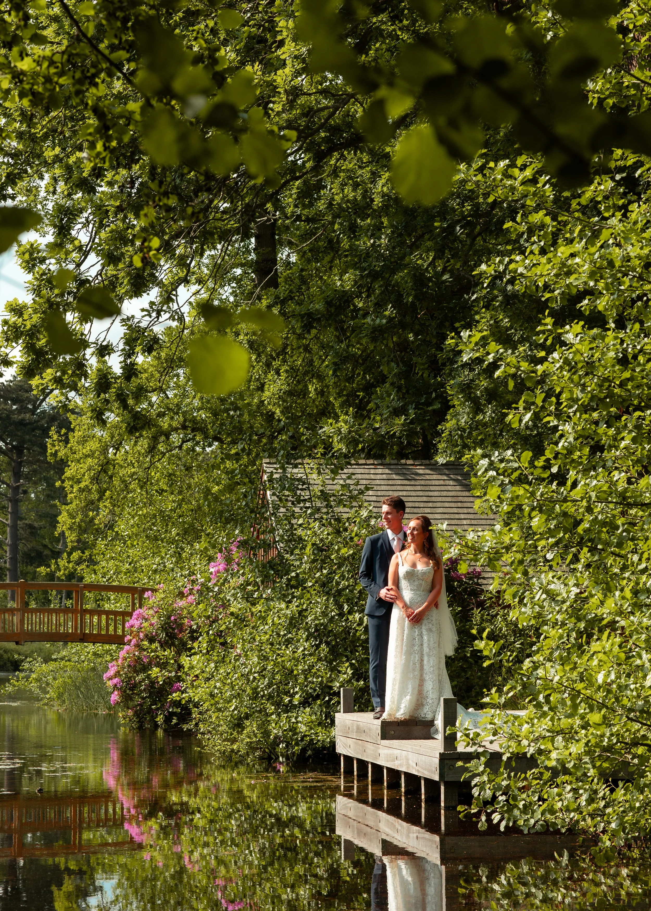 Bride and groom standing on a wooden dock beside a pond, surrounded by lush green trees and flowering bushes with pink flowers, under a bright blue sky.