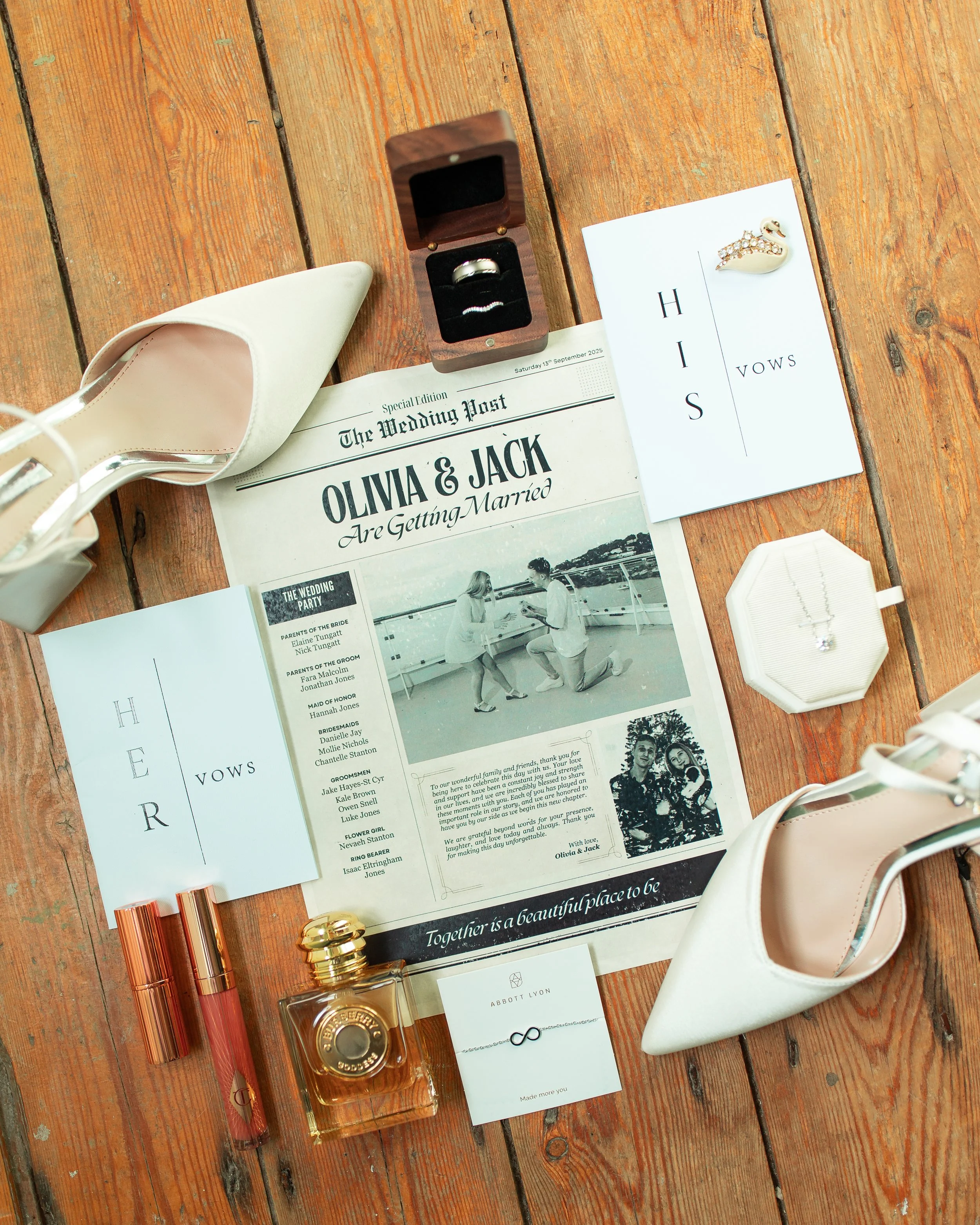 Wedding accessories and printout on wooden surface, including white high-heeled shoes, gold perfume, lipstick, wedding rings in wooden box, vows cards, swan brooch, necklace in case, and wedding announcement for Olivia and Jack.