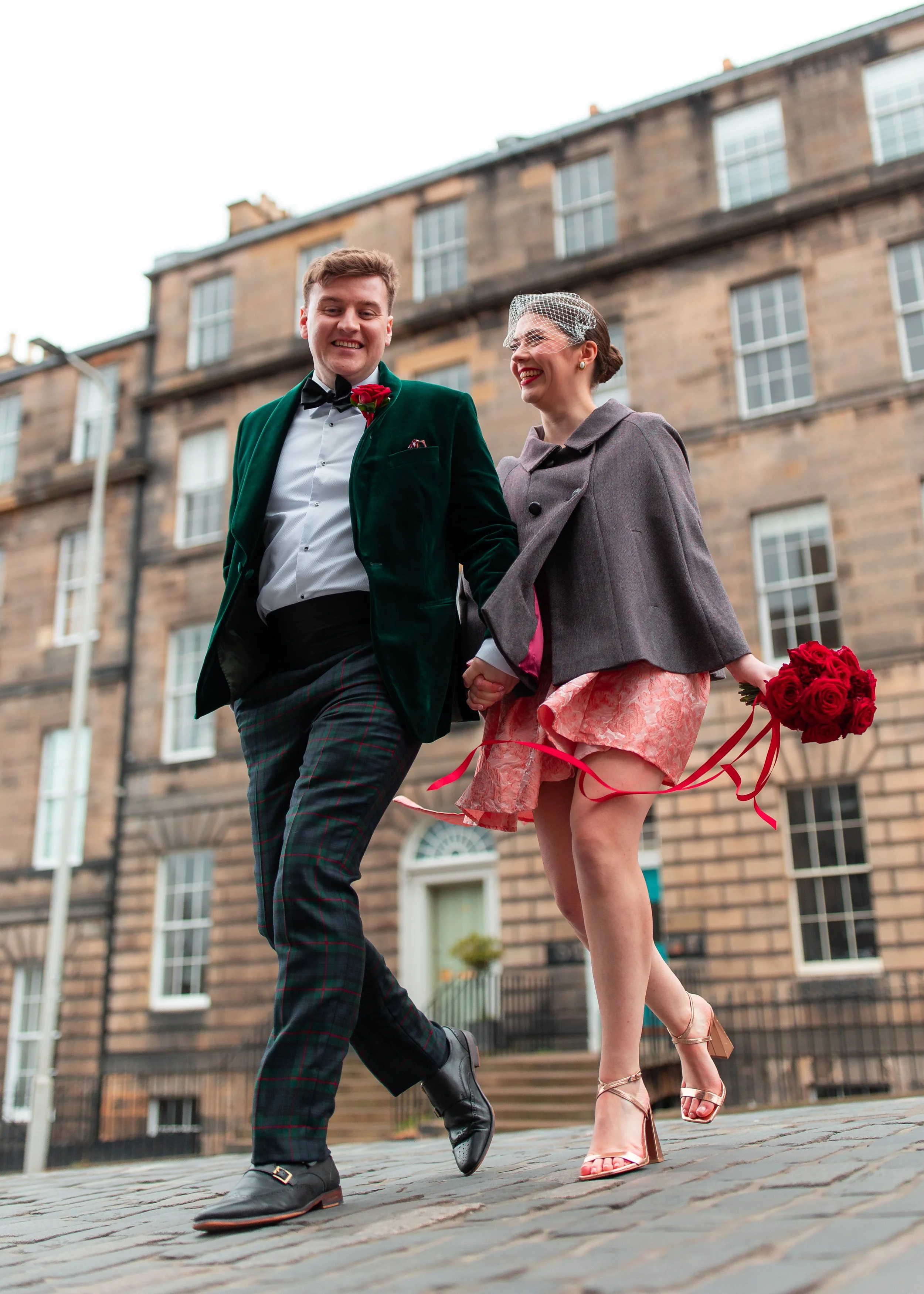 A couple dressed in vintage clothing, holding hands and smiling as they walk on a cobblestone street in front of a historic brick building. The woman is carrying a bouquet of red roses and wearing high heels, while the man is wearing a green velvet b