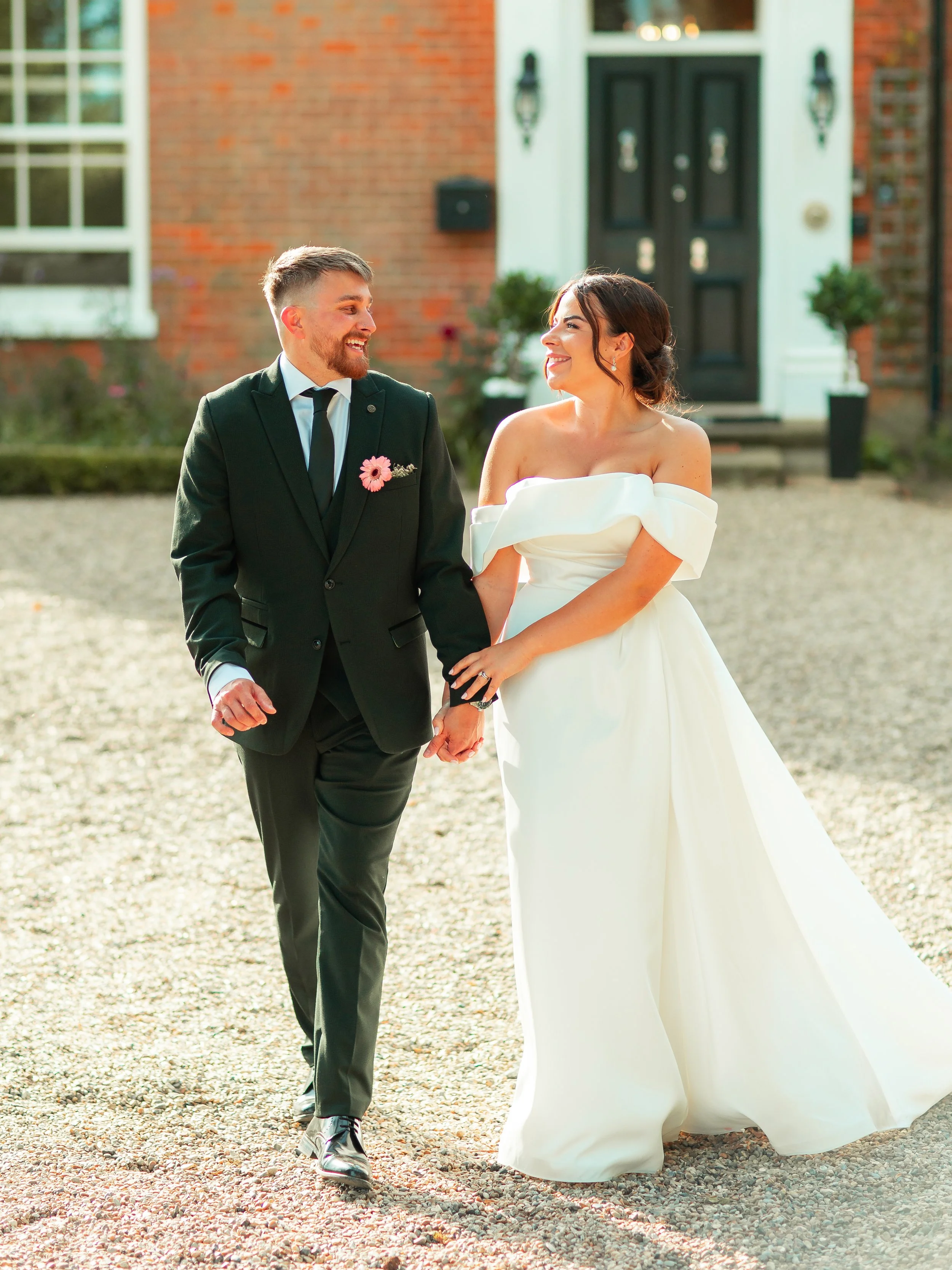 A happy bride and groom holding hands and walking outside in front of a brick house, smiling at each other on their wedding day.