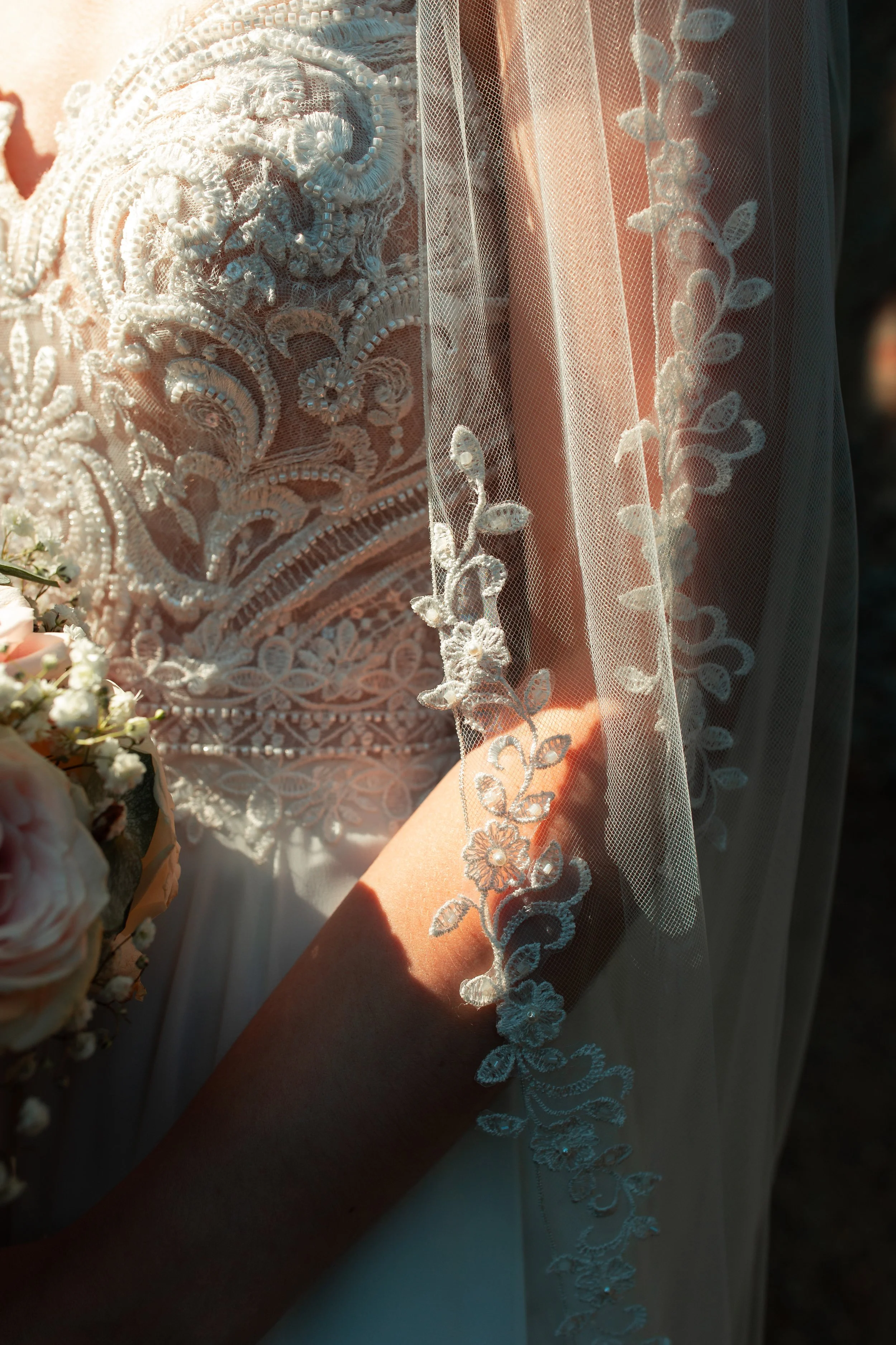 Close-up of a bride's lace wedding dress with intricate embroidery, holding a bouquet of flowers.
