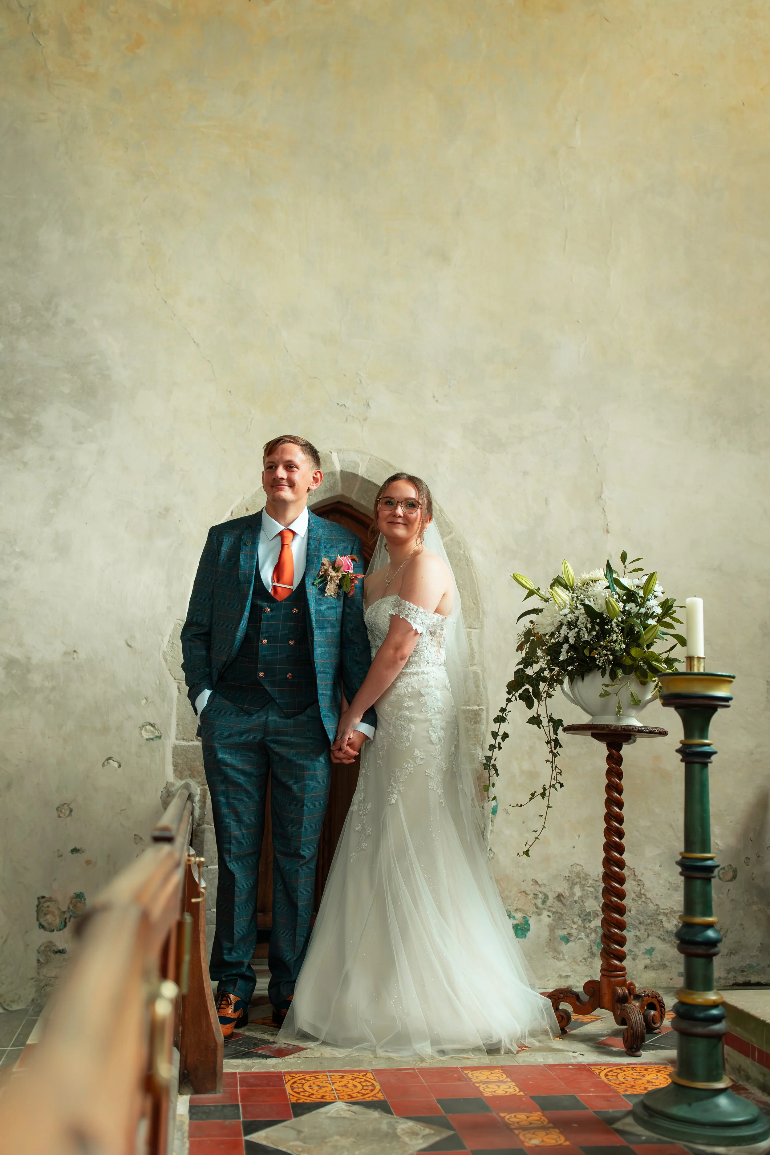 A bride and groom holding hands during their wedding ceremony inside a church or chapel, standing close in front of a textured, rustic wall near an archway, with a flower arrangement on a pedestal and a candle nearby.
