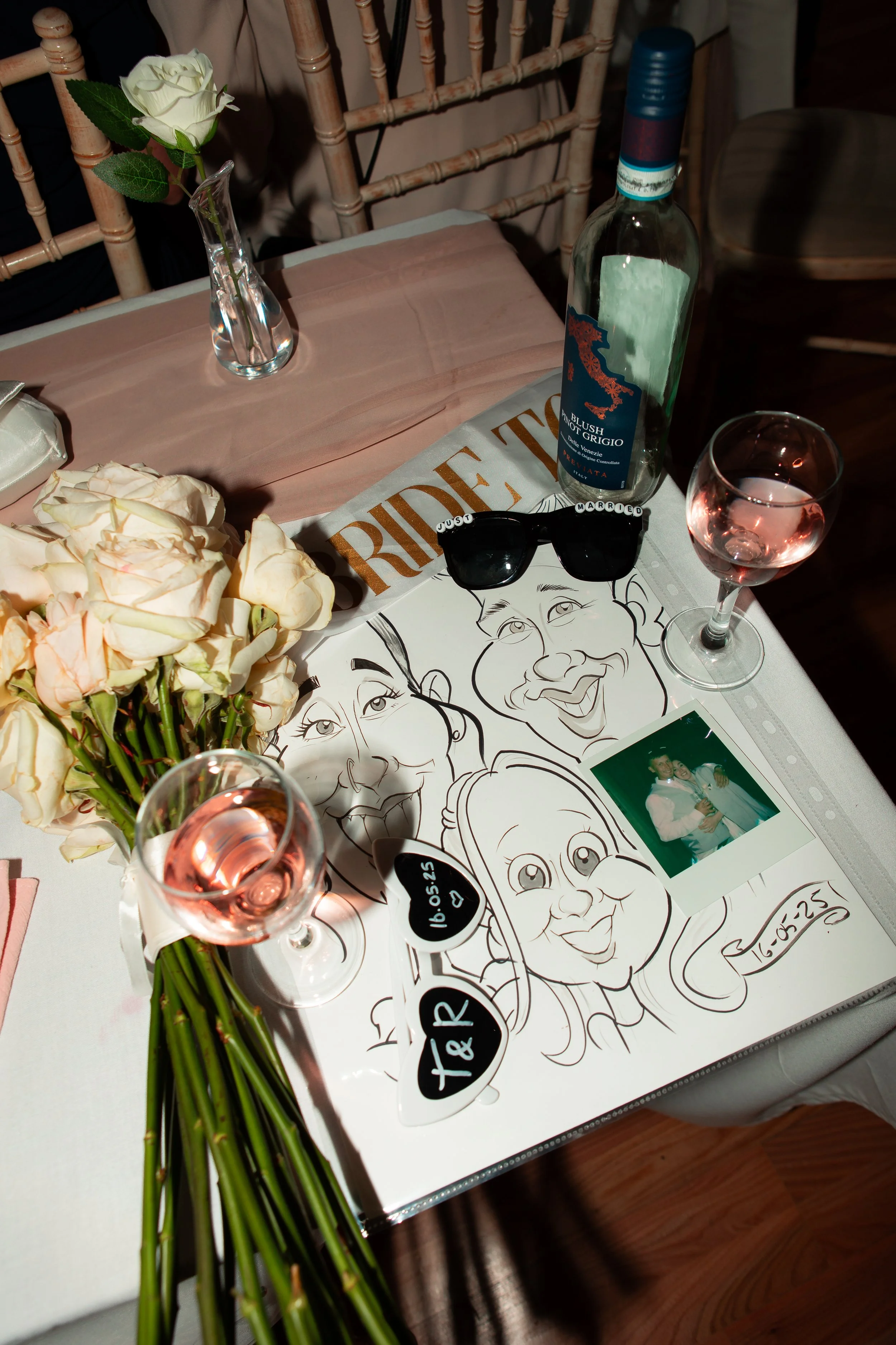 A table setting with a vase of white roses, a bottle of rosé wine, two glasses of rosé wine, black sunglasses, a caricature sketch of two people, a printed photo of two people, and decorative signs with the date 16.05.25. The table has a pink tablecl