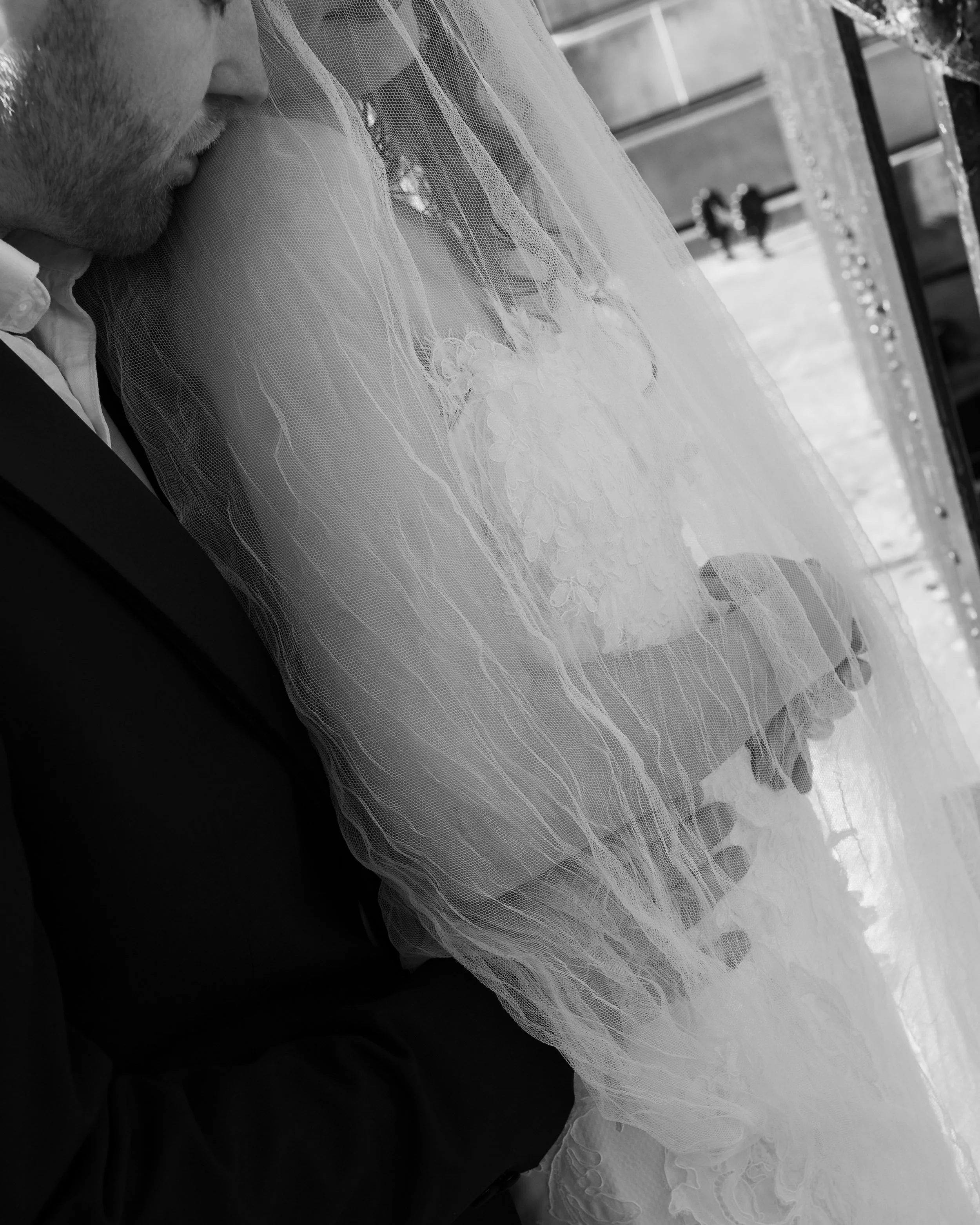 Close-up of a bride in a wedding gown with lace details, standing next to a person in a dark suit, both holding hands, with a veil partially covering the scene.