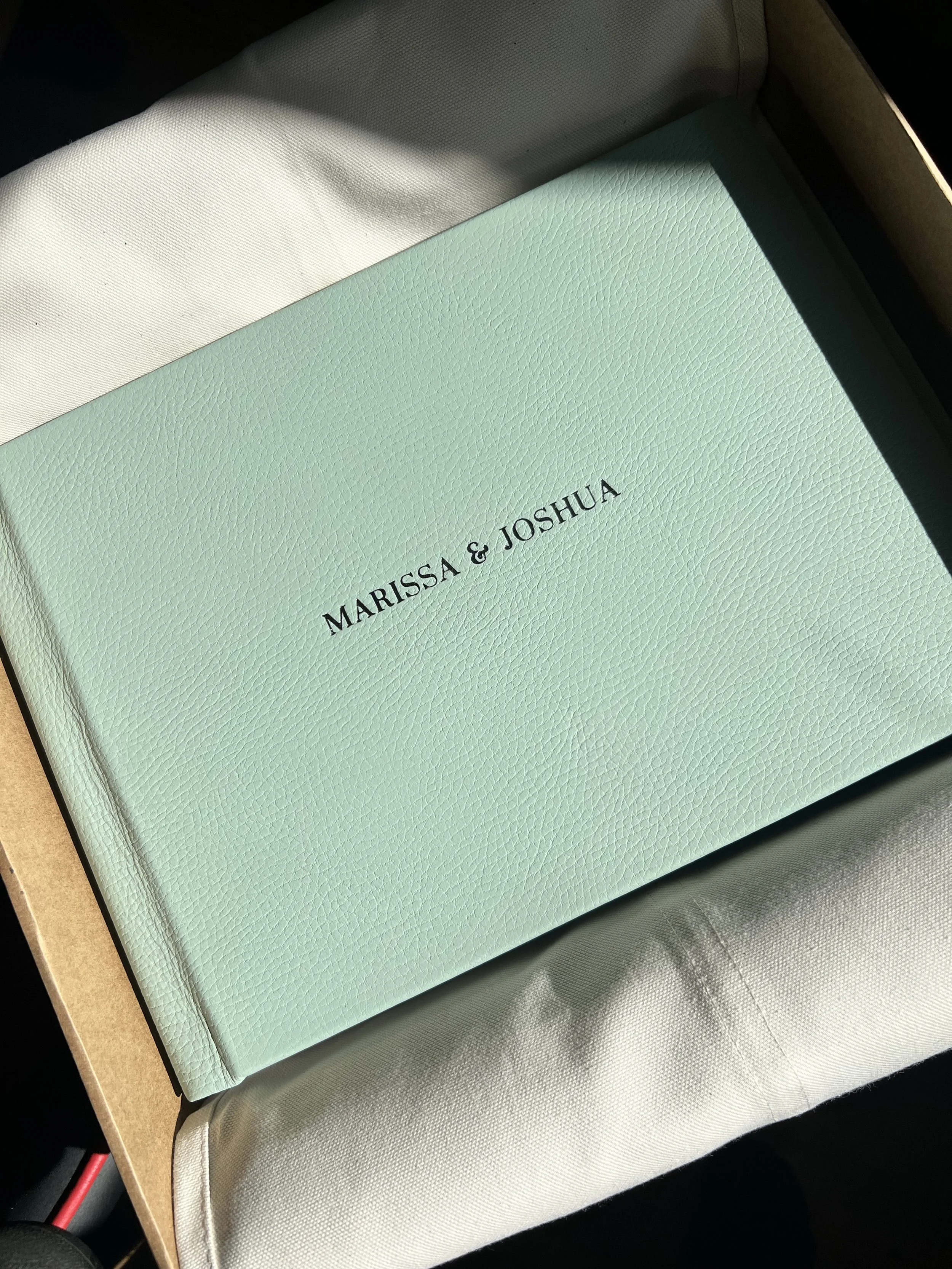 A light green wedding album, with the names 'Marissa & Joshua' printed in black, resting on white tissue paper inside a cardboard box.