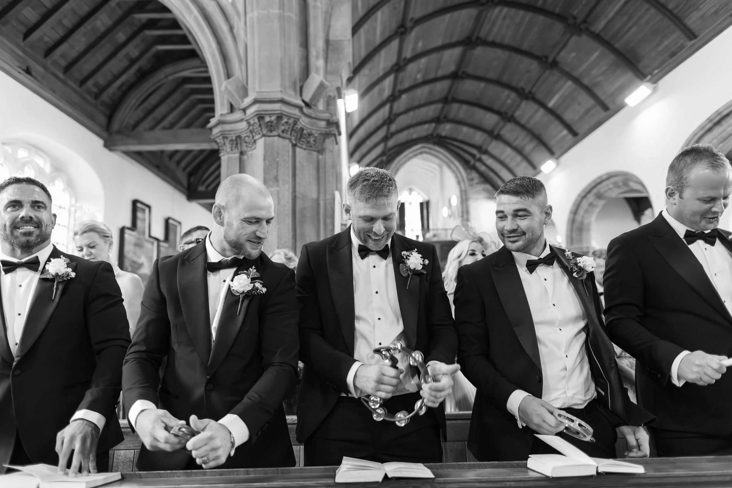 Groom and groomsmen at a wedding ceremony, holding tambourines inside a church with wooden ceilings.