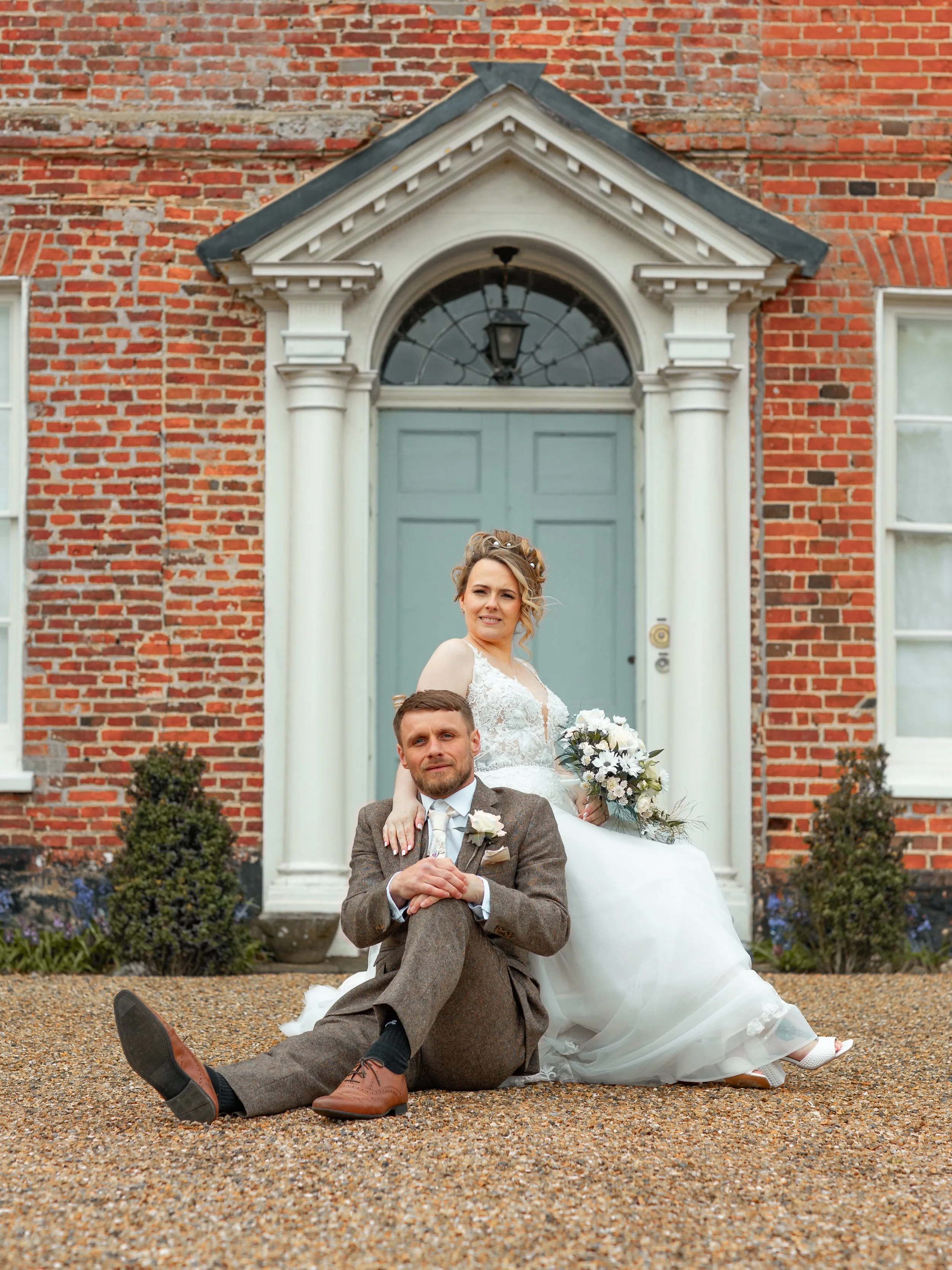 A bride and groom sitting and leaning against each other in front of a brick house with white columns and a blue door. The bride is wearing a white wedding dress and holding a bouquet, while the groom is dressed in a brown suit with a white shirt and a tie.