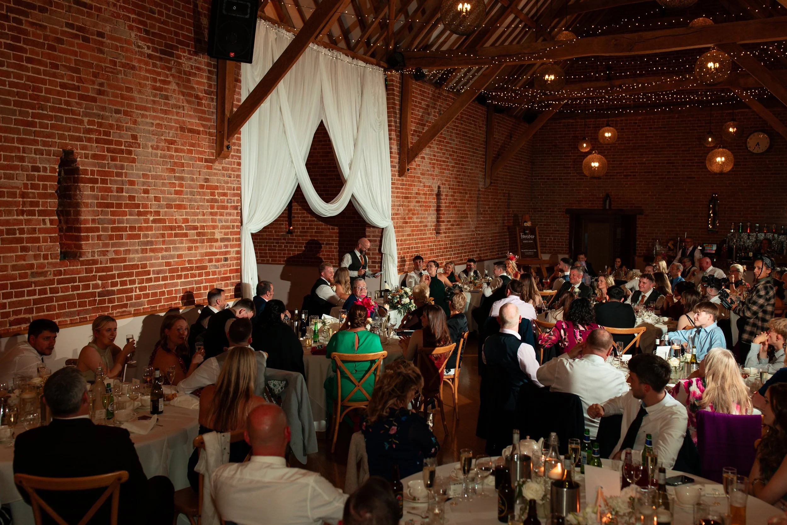 Guests seated at round tables in a decorated banquet hall with exposed brick walls, string lights, and white drapery, listening to a speaker at the front.