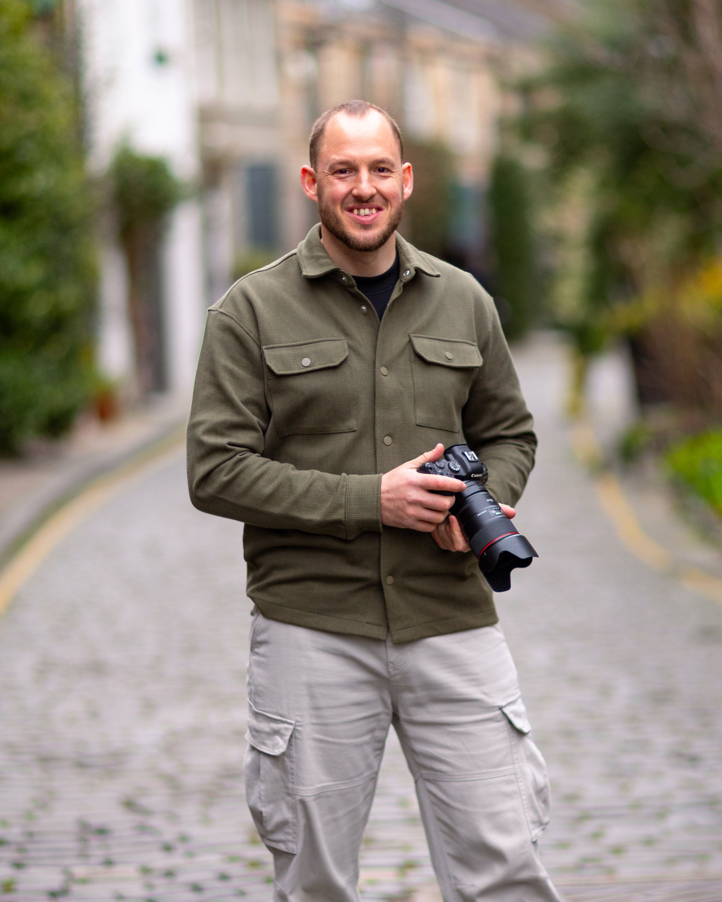 A man holding a camera, standing on a cobblestone street with blurred greenery and buildings in the background.